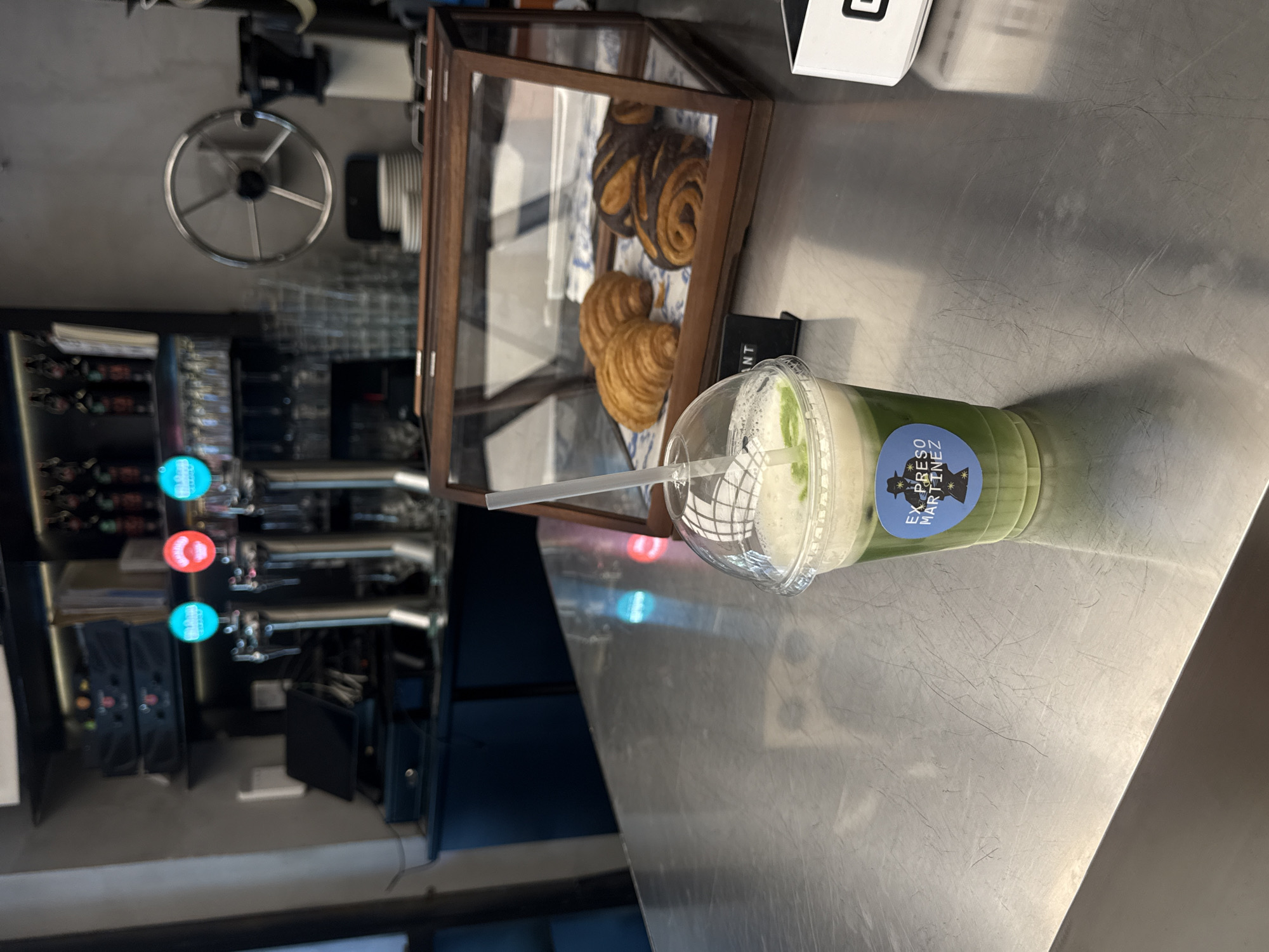 The image depicts a counter in what appears to be a café or bakery. On the counter is a plastic cup filled with a green liquid, likely a matcha latte, with a straw. The cup has a label that reads "Expreso Martinez," suggesting the location is a café or bakery of that name.
Next to the cup is a wooden-framed display case containing pastries, specifically croissants and pain au chocolat. In the background, a soda machine and a metal-framed object are visible, indicating a food service environment. The countertop is stainless steel.
The café "Expreso Martinez" is located in Madrid, Spain.