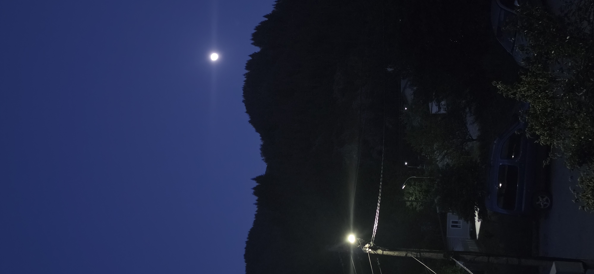 The image depicts a night scene in what appears to be a residential area. A dark silhouette of a mountain rises along the center of the frame, with a bright full moon positioned over it in the dark blue sky. A street light illuminates the bottom right portion of the image, revealing power lines, a building, foliage, and a vehicle. Given the prompt, the location is likely Rudozem, Bulgaria.
