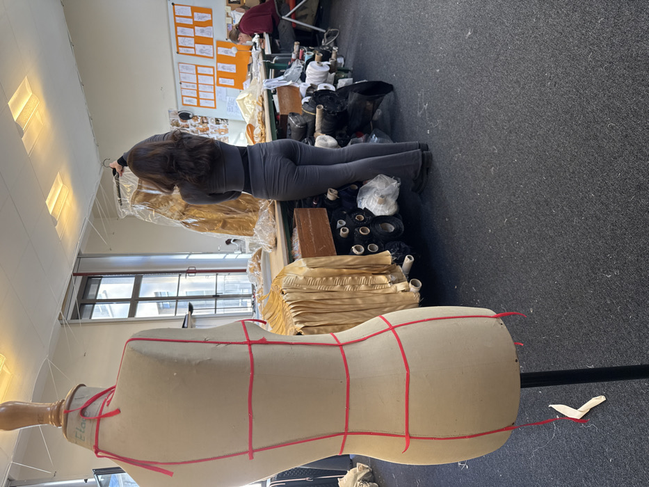 The image displays an indoor fashion design or textile workshop located in Arcueil, France. In the foreground, a cream-colored female dress form, marked with red ribbon strips indicating design lines, stands on a black pole base, positioned with its back towards the viewer. Behind it, a female individual, seen from the back, with long dark brown hair, wears a dark gray long-sleeved top and matching flared trousers. She is holding a golden-yellow garment, contained within a clear plastic garment bag, by its hanger, elevated above her head.
A large work table occupies the midground, featuring draped golden-yellow fabric, some exhibiting pleated textures. Beneath this table, numerous rolls of various fabrics, including black and white materials, are stored on shelving or directly on the dark gray carpeted floor. In the background right, a second individual with lighter hair, wearing a dark reddish-brown top, is seated at a table, concentrating on materials in front of them. The back wall features a display board adorned with multiple sketches or design drafts on white paper, some with orange backings. Large windows on the left wall provide natural light, revealing an exterior building with architectural details. Square, recessed ceiling lights are visible, and thin white strings are strung horizontally across the upper section of the room. The floor is covered with dark gray carpeting, showing scattered fibers. A small piece of white material rests on the floor near the base of the dress form.