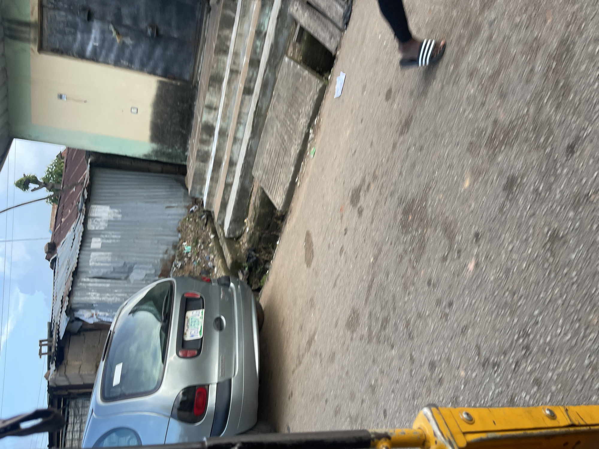 The image depicts a street scene, likely in Jos, Nigeria, with a silver minivan parked on the right side of the road. In the foreground, a portion of a yellow vehicle, possibly a Keke Napep (a three-wheeled auto-rickshaw commonly used as a taxi), is visible on the left. The background shows a row of buildings with corrugated iron roofing, some appearing weathered and dilapidated. Adjacent to the minivan are concrete steps leading up to a building with a yellow and green facade and a dark door. The ground is paved with asphalt and shows signs of wear and scattered debris. A person's legs and feet, wearing black pants and sandals, are visible walking on the right side of the road. The sky is partly cloudy. The overall impression is one of a densely populated urban environment with signs of both commercial activity and potentially challenging living conditions.