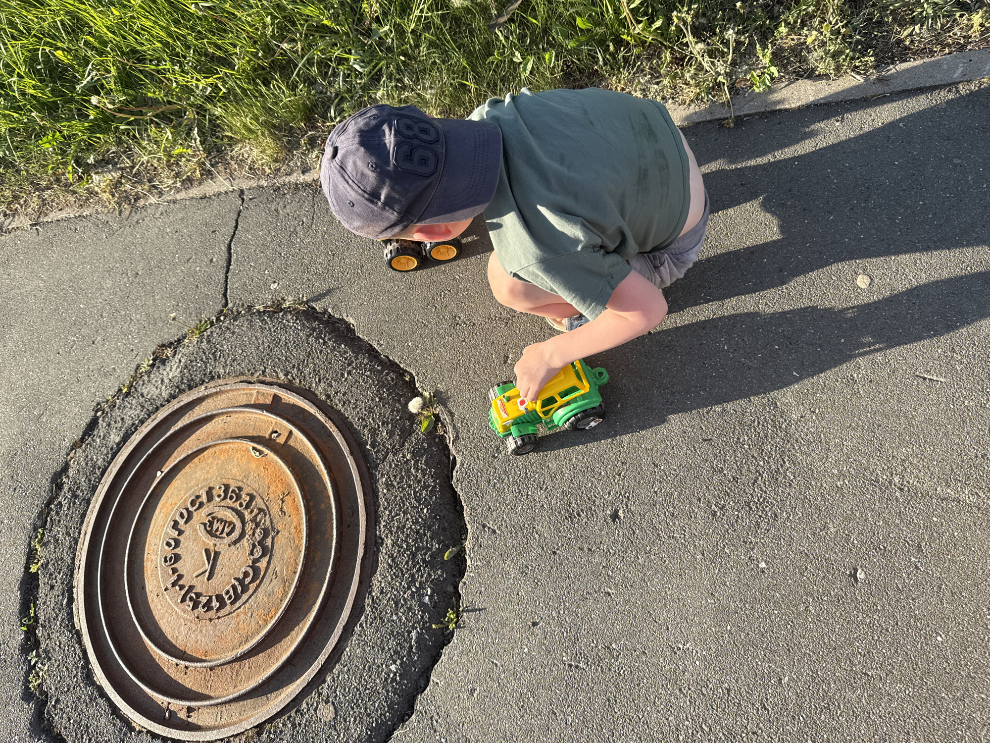 A child is playing with toy vehicles near a manhole cover on an asphalt surface. The child, wearing a blue cap with the number "8" on it, a green shirt, and grey shorts, is crouching and appears to be positioning a yellow and green toy truck. Another toy vehicle, with yellow wheels, is visible near the child's legs. The manhole cover is round and rusty, with raised concentric rings and stamped text in Russian, identifying the location as the city of Прикамский, Russia. The scene is partially illuminated by sunlight, casting shadows of the child and the objects on the pavement. The edge of a grassy area is visible in the upper left corner of the image. The surface of the pavement has cracks and minor vegetation growing in the gaps.