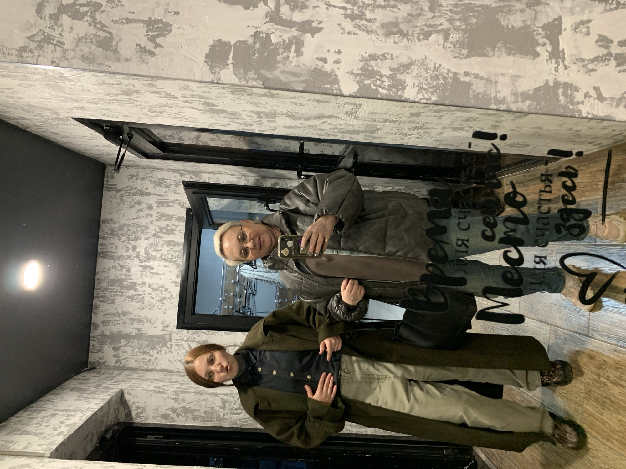 Two women are captured in a mirror selfie indoors, likely in Chaikovsky, Russia, given the location context and visible Russian text. The older woman, with short blonde hair and glasses, wears a grey padded jacket and is holding a smartphone to take the picture. She has rings on her fingers and light-colored nail polish. The younger woman stands in front, with brown hair, dressed in a long dark green coat, a dark top with light buttons, and light khaki trousers. She is carrying a small black handbag and is wearing patterned shoes. Both women have neutral to slightly smiling expressions.
The setting appears to be a room or hallway with a textured wall treatment, featuring a distressed pattern of light grey and white. A dark grey wall section is visible on the left, with a single wall light fixture. The floor is made of warm-toned wooden planks. The image is taken in a large mirror, which reflects the room and the people. The mirror itself has dark frames, suggesting it might be part of a larger decorative element or a series of framed mirrors/windows.
A phrase written in a calligraphic style is visible on the mirror, appearing reversed in the original image. When read correctly (as if viewing the actual mirror), the Russian text states: "Момент счастья здесь!", which translates to "A moment of happiness is here!". The indoor lighting suggests it is not daytime or that the windows do not offer much natural light.