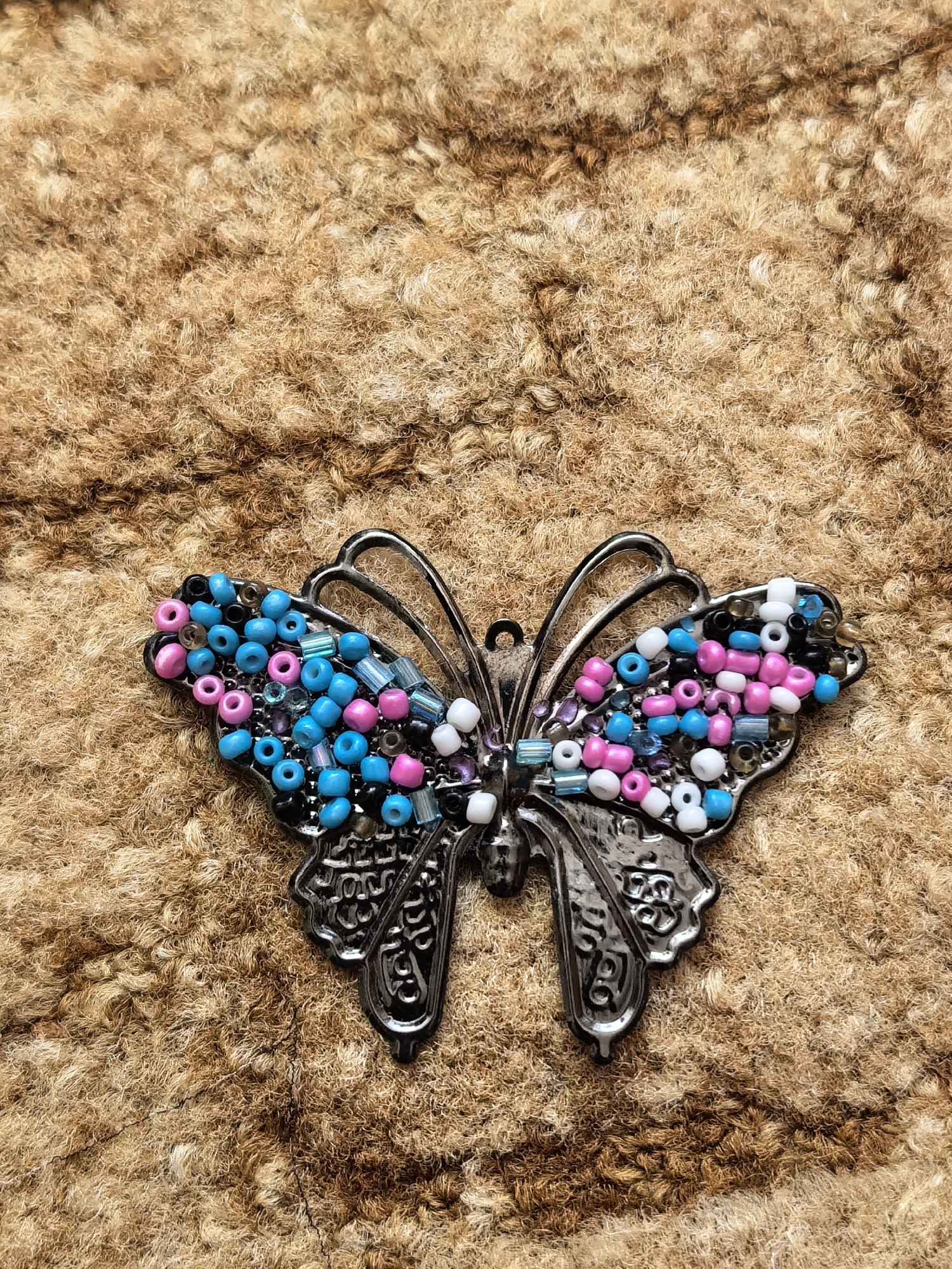 A butterfly-shaped brooch lies on a textured, brown surface. The brooch has a silver-toned metal frame decorated with small, colorful beads in shades of blue, pink, white, and black. The beads are attached to the upper part of the wings. The metal frame has ornate details and shapes resembling butterfly wings. The textured surface in the background appears to be a close-up of a material like wool or fur. The image does not provide information about Sterlitamak, Russia, so no details can be provided regarding this city.