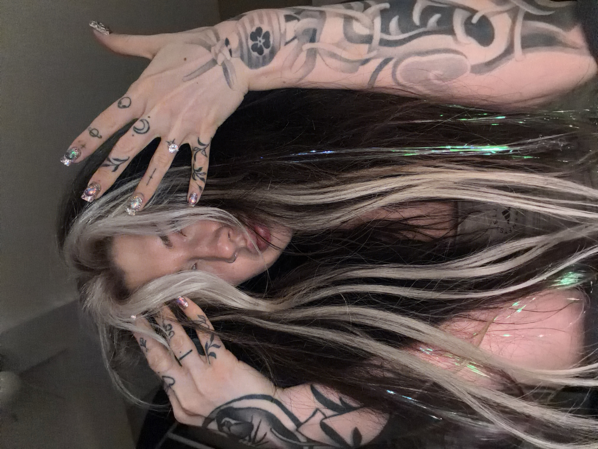 A young individual with fair skin and dark brown hair with blonde streaks is captured from an unusual perspective, seemingly an overhead shot taken at an angle. The person's hair is long, flowing across the frame, with glittery hair tinsel adding a sparkling effect. Their hands, heavily tattooed with black ink designs, are raised and positioned near their face. The tattoos on their hands include celestial symbols like a crescent moon and stars, along with floral and abstract patterns. Their fingernails are adorned with iridescent designs. The person's face is partially obscured by their hair and hands, but their lips are visible, and a small silver hoop earring is noticeable in one ear. The background is dimly lit, suggesting an indoor setting, with indistinct details suggesting an interior space. The location of Budva, Montenegro is not discernable from the image.