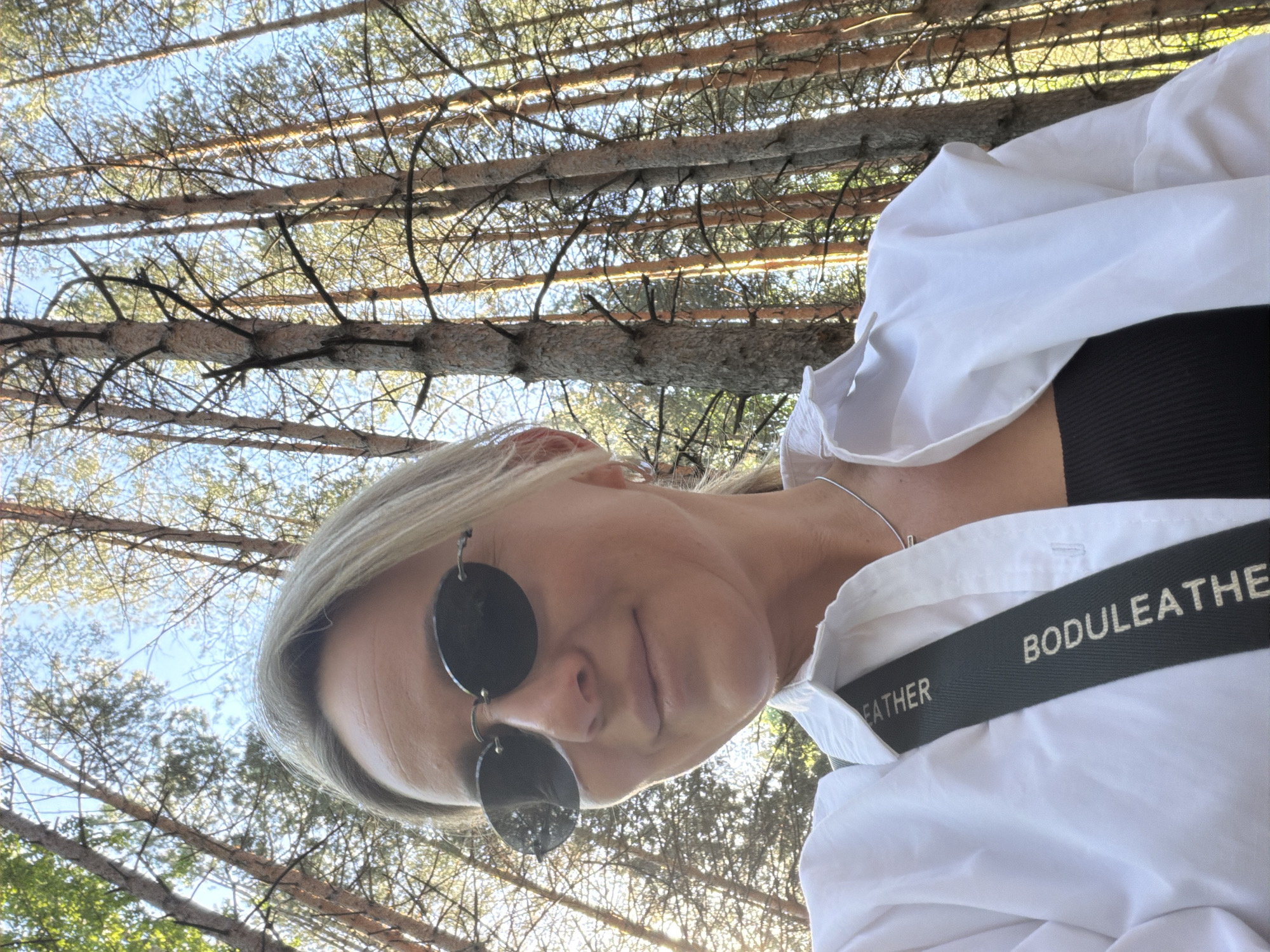 A person is captured in a selfie taken from a low angle, looking upwards. The person is wearing sunglasses with dark, circular lenses and a white collared shirt over a black top. A strap or accessory with the inscription "BODULEATHER" is visible on their shoulder. The background consists of tall, slender trees, possibly in a forest or wooded area. The angle of the shot and the presence of trees suggest the location is outdoors. The scene is lit by natural sunlight, which filters through the trees. There is no clear evidence to confirm the exact location in the city of Chaikovsky, Russia.