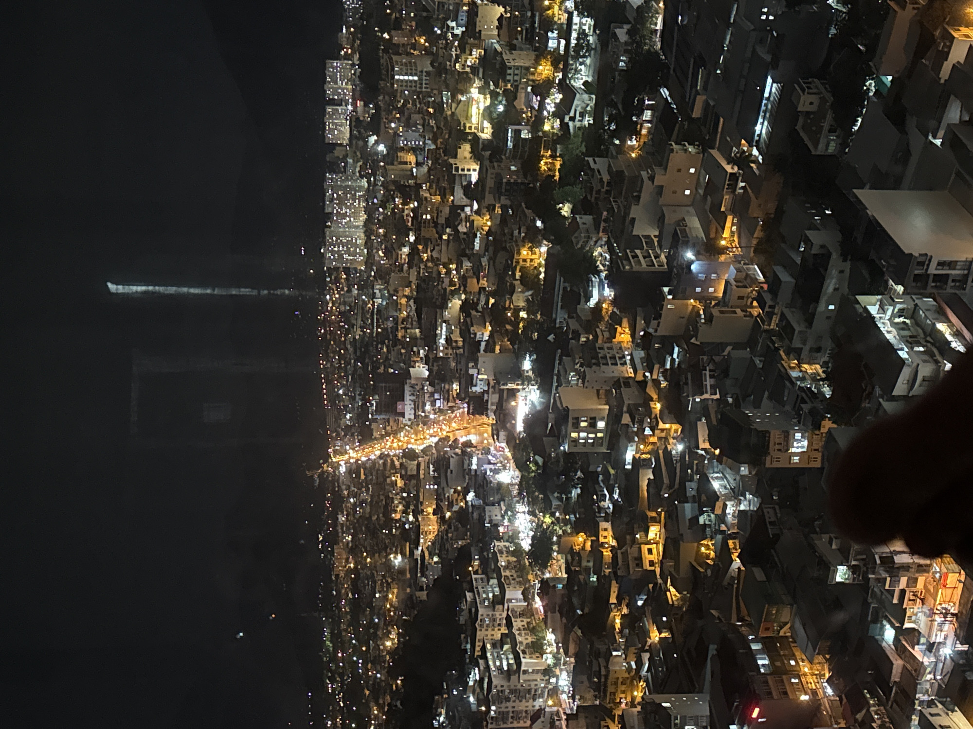 This is a nighttime aerial photograph of a densely populated city with numerous buildings and illuminated streets. The city is sprawling, with lights visible in many windows and along roads, suggesting it is a busy urban area. In the distance, taller buildings, possibly apartments or hotels, are brightly lit. A prominent road, illuminated by streetlights and vehicle headlights, cuts through the cityscape, indicating a main thoroughfare. The overall scene suggests a vibrant city at night, likely in a tropical or subtropical region given the dark, clear sky.
While the exact location is not definitively identifiable from the image alone, the presence of a vibrant, busy city with a coastal feel and the specific architectural styles of the buildings are consistent with Nha Trang, Vietnam. Nha Trang is known for its long sandy beaches, bustling city center, and growing skyline of hotels and apartments. The dense urban development, the illuminated main street, and the mountainous backdrop visible in some parts of the image align with common views of Nha Trang.