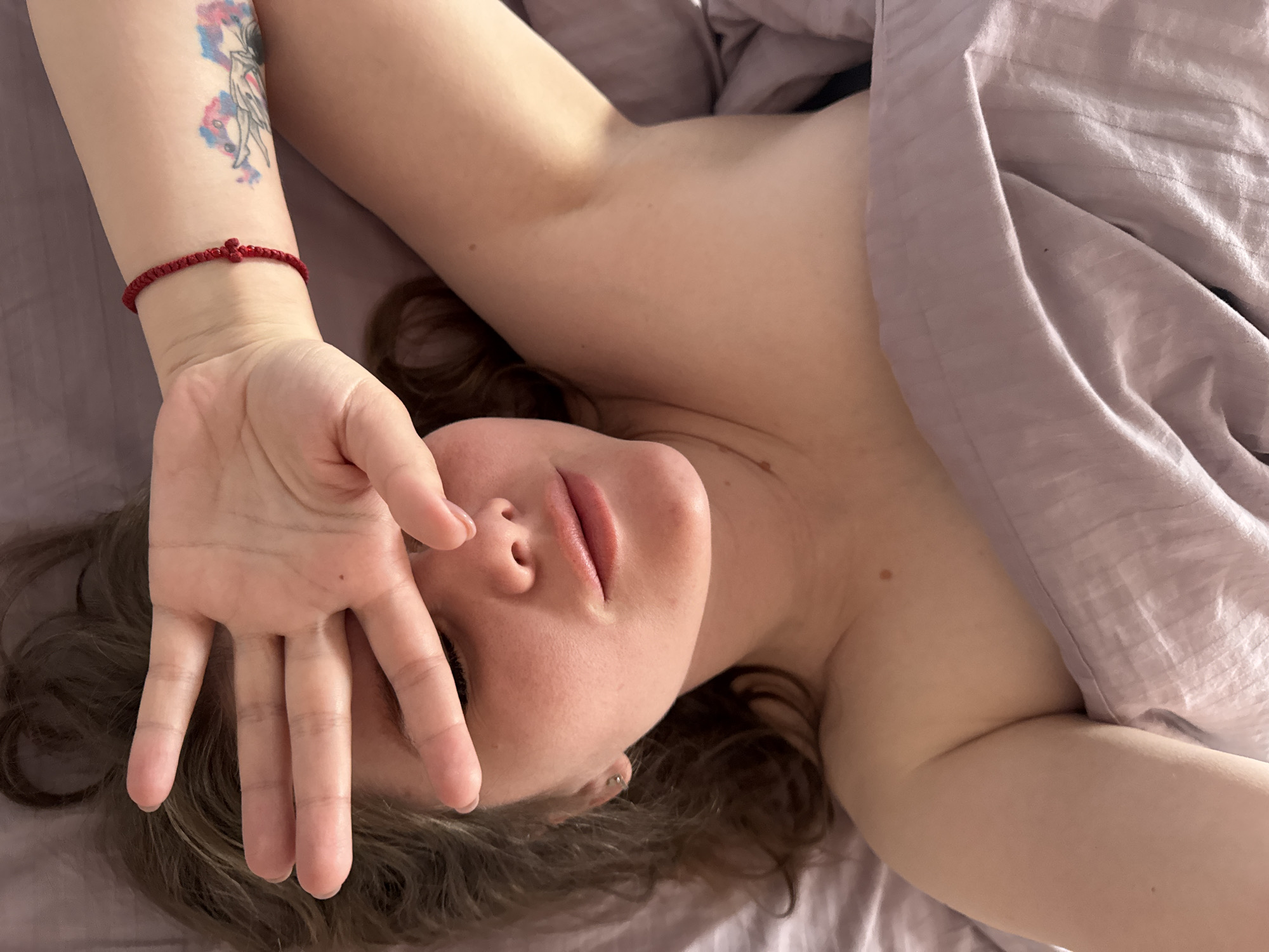 A fair-skinned woman lies on a lavender bed, partially covered by a matching lavender blanket. Her right arm is extended above her head, the hand partially obscuring her face. A red woven bracelet is visible on her right wrist, and a colorful tattoo is present on her right forearm. Her left arm is positioned beside her. She has medium-length brown hair and a visible earring in her left ear. The scene is lit with natural light. The location is unspecified, and there is no identifiable information to confirm the location in Gornyi, Dvurechenskoe S/P, Russia.