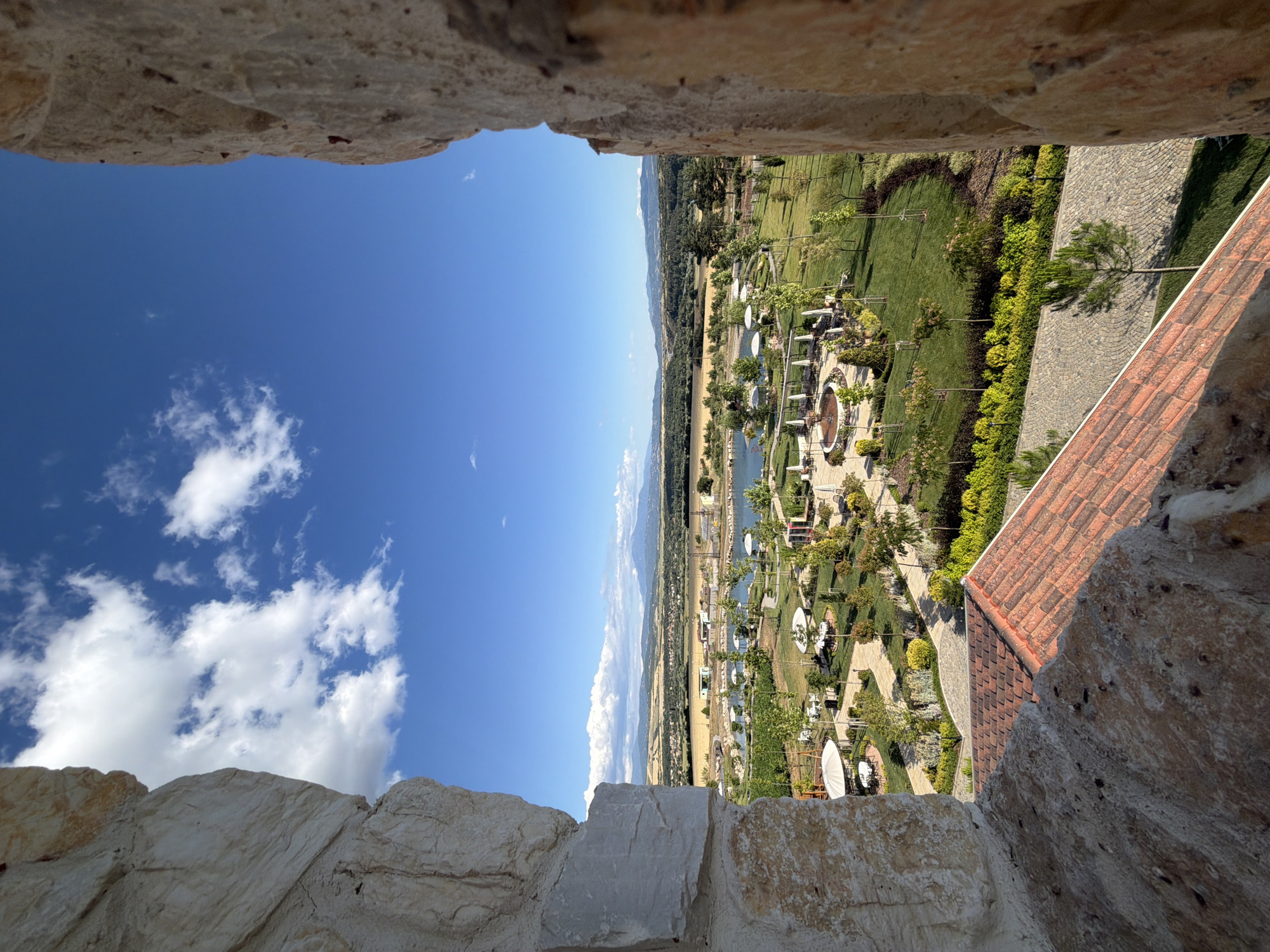 The image is a vertical, eye-level, outdoor shot captured from a high vantage point, likely a castle window or parapet, looking out over a landscaped park and surrounding countryside. The foreground features rough, textured stone walls framing the view on the left and right, and a portion of a terracotta tiled roof in the lower foreground.
Beyond the stone structures, the scene opens up to a well-maintained park with lush green lawns, mature trees, and decorative shrubs. Several circular seating areas with white umbrellas and tables are scattered throughout the park. A body of water, possibly a pond or small lake, is visible in the mid-ground, reflecting the blue sky. In the distance, the landscape transitions to rolling hills covered in fields of golden crops and interspersed with patches of forest. Further back, a range of mountains forms the horizon, under a bright blue sky dotted with fluffy white clouds. The overall impression is one of a serene and picturesque rural setting.
The scene is likely located in the vicinity of Kocherinovo, Bulgaria, given the architectural style of the stone structures and the type of rural landscape depicted. There are no people visible in the image. The time of day appears to be daytime, with clear weather conditions.