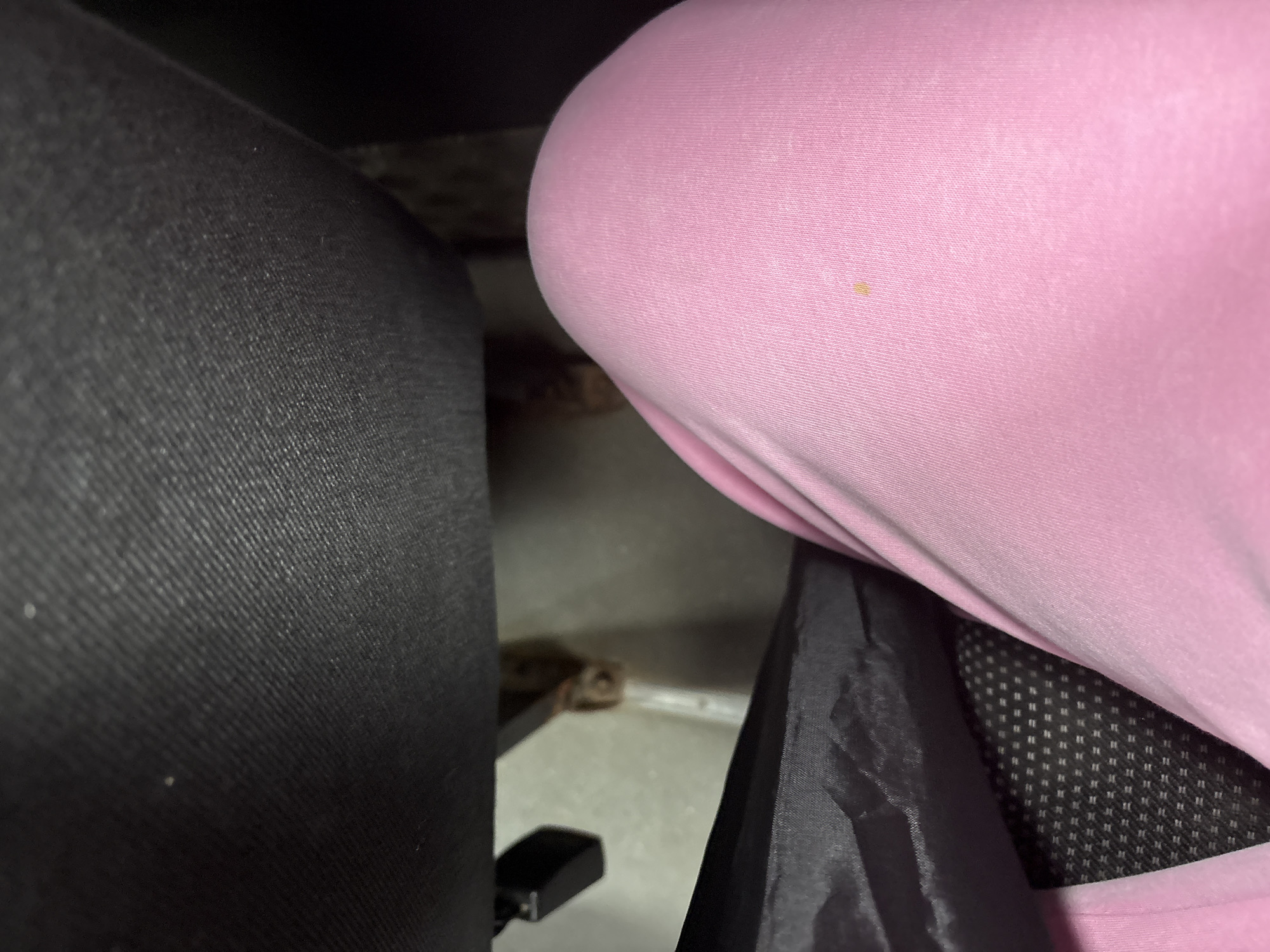 The image provides an upward-angle, close-up view of a person's legs wearing pink pants. The pants have a small yellow stain on the right thigh. To the left of the legs, a black umbrella is visible, partially obscuring a dark-colored textured fabric, likely part of a chair. In the background, there is a light-colored, concrete-like surface with metallic elements, suggesting an indoor or possibly an underpass setting. There are no visible identifying features of Mahilyow, Belarus, within the frame. The overall scene is domestic or personal, focusing on clothing and personal accessories.