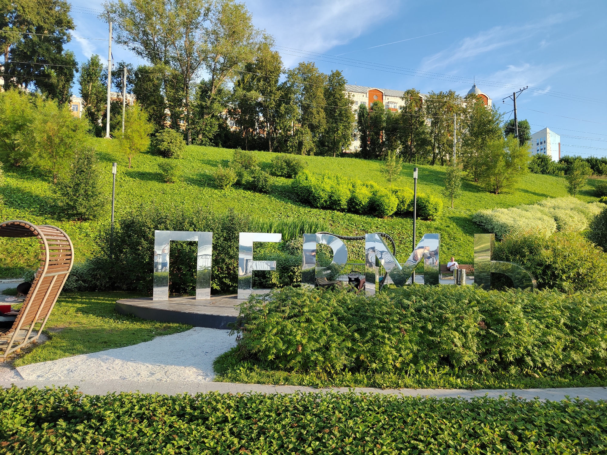 The image depicts a public park in the city of Perm, Russia, featuring large, reflective, three-dimensional metallic letters spelling out "ПЕРМЬ" (Perm). The letters are positioned in the foreground, partially obscured by lush green bushes. Behind the letters, a gently sloping grassy hill is adorned with various trees and shrubs, and residential buildings are visible in the background. To the left, a curved wooden bench-like structure is present. The sky is blue with scattered clouds. In the middle ground, a person is sitting on a bench behind the letters, reading a book. The scene is bathed in natural sunlight, indicating it was taken during the daytime.