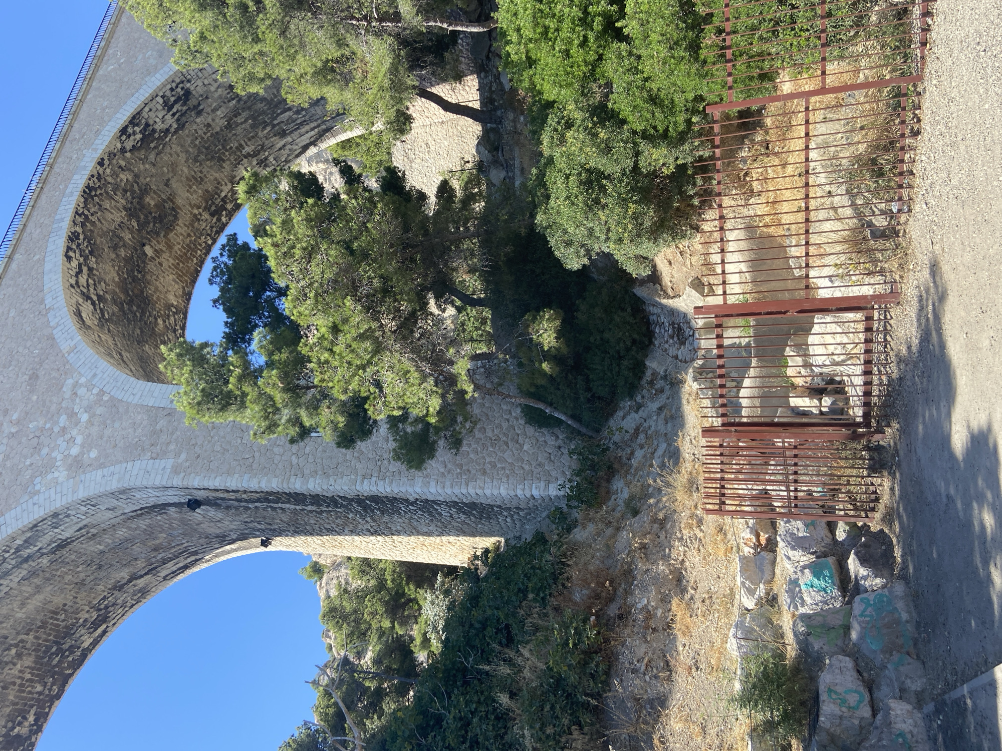 A low-angle, eye-level outdoor shot shows a large stone bridge arching over a rocky, vegetated slope on a sunny day. The bridge, constructed of large, light-colored stones with some darker, weathered stones visible underneath its arch, dominates the upper portion of the frame. The sky is a clear, bright blue.
The slope beneath the bridge is covered in a mix of green bushes, dry grasses, and large rocks, some of which have graffiti in teal. A reddish-brown metal gate, partially open, stands in the foreground, partially obscuring the view of the rocky terrain behind it. Several trees with green foliage grow on the slope, some appearing to be pine trees. The ground in the immediate foreground is a gravel path.