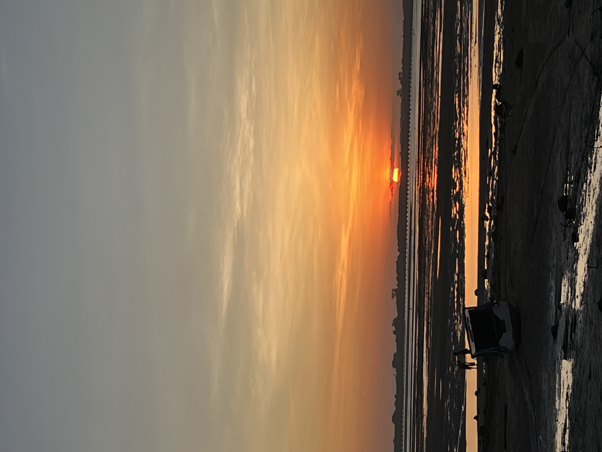 This image captures a serene sunset scene over what appears to be extensive tidal flats or a shallow bay, located in Kuta Selatan, Indonesia.
The sky dominates the upper portion of the frame, displaying a gradient of colors from a light, muted grey-blue on the left to vibrant oranges and yellows towards the setting sun on the right. Streaks of wispy clouds are visible, catching the warm light.
The sun, a brilliant orange orb, is positioned low on the horizon, partially obscured by a thin, dark silhouette of what could be a distant landmass or a man-made structure, possibly a bridge. Its intense light casts a warm glow across the entire sky.
The middle ground and foreground consist of dark, textured mudflats or very shallow water. These surfaces strongly reflect the sunset, creating elongated, shimmering vertical streaks of orange and yellow light that contrast with the darker areas.
On the far right in the foreground, a small, dark, box-like structure, possibly a boat or a basic shelter, is visible on the dark ground, appearing as a silhouette. Irregular dark shapes, likely rocks or debris, are also scattered across the dark shore.
The scene conveys a peaceful atmosphere at dusk. There are no visible people or discernible text in the image.