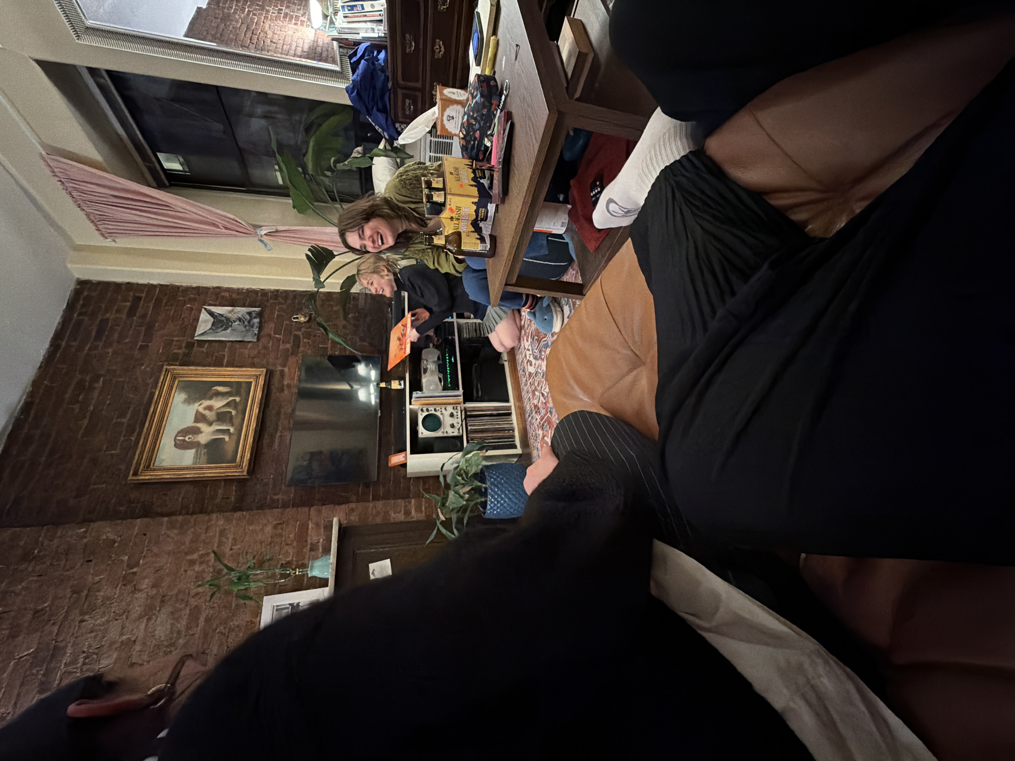 The image shows an interior scene likely in a residential space in New York City, United States. A group of people are situated around a coffee table. One person is holding a vinyl record, while another is laughing. There are bottles of beer on the table. The room features exposed brick walls, artwork including a framed painting of a dog, a television, and shelves containing various items. A window with a pink valance is visible. The focus is partially obscured by the photographer's position.