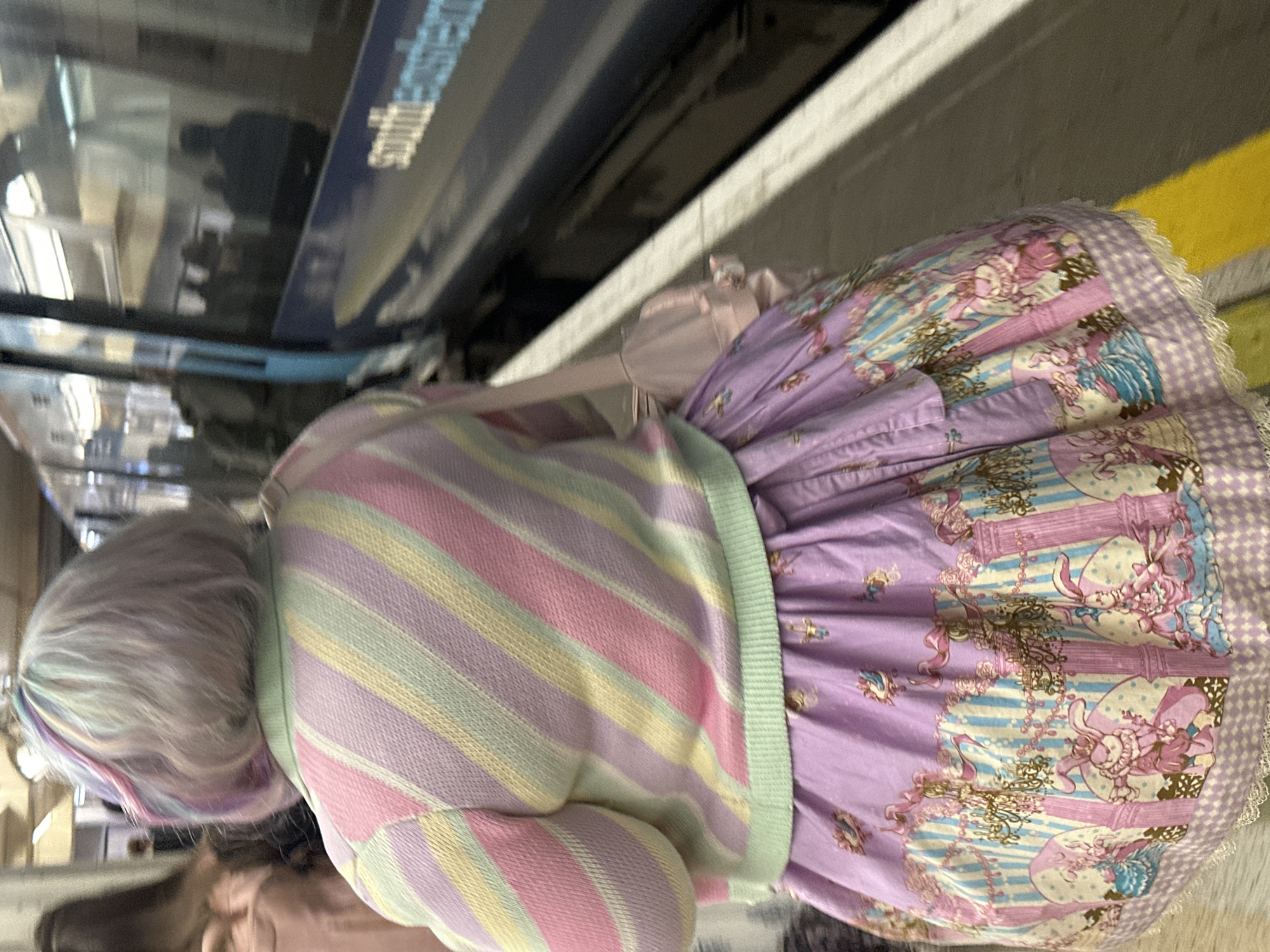 A person in pastel clothing stands on a train platform next to a Southeastern train in London, UK. The individual wears a striped sweater in pastel shades of pink, yellow, green, and lavender, and a frilly lavender skirt with a print. The person's hair is a light gray color with streaks of blue and purple. The train is blue with the word "Southeastern" visible on its side. A yellow line is marked on the edge of the platform.