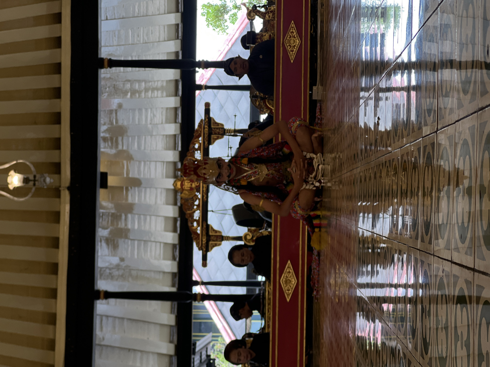 The image depicts a cultural performance in a large, open pavilion with a striped canopy and ornate wooden structures. In the foreground, a young person dressed in traditional Javanese attire, including a crown and elaborate makeup, is seated cross-legged on a patterned tiled floor. The performer is adorned with necklaces and a patterned cloth wrapped around their waist. Behind the performer, a group of individuals are seated in what appears to be a musical ensemble, possibly playing gamelan instruments which are partially visible. They are dressed in black attire, with the men wearing traditional Javanese headwear. The setting suggests a formal cultural event, possibly a traditional dance or music performance, likely taking place in a historically significant location in Yogyakarta, Indonesia, such as a palace (kraton) or a cultural heritage site. The polished tiled floor reflects the overhead structures and the performance, adding to the visual richness of the scene.