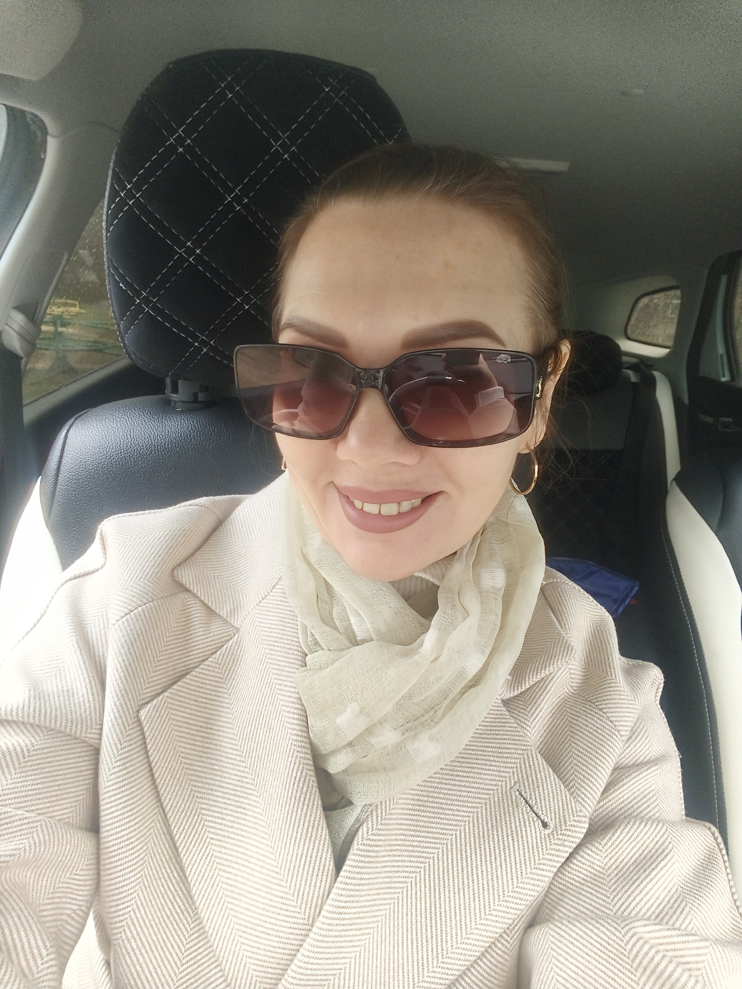 A woman is photographed in the front seat of a car. She is wearing large, rectangular sunglasses, gold hoop earrings, a cream-colored scarf, and a blazer with a herringbone pattern. The car's interior features black and white leather seats with a quilted headrest. The car appears to be parked, and outside the car window, there is a glimpse of trees and greenery. The image suggests the location is in a car but provides no indication of the city or region.