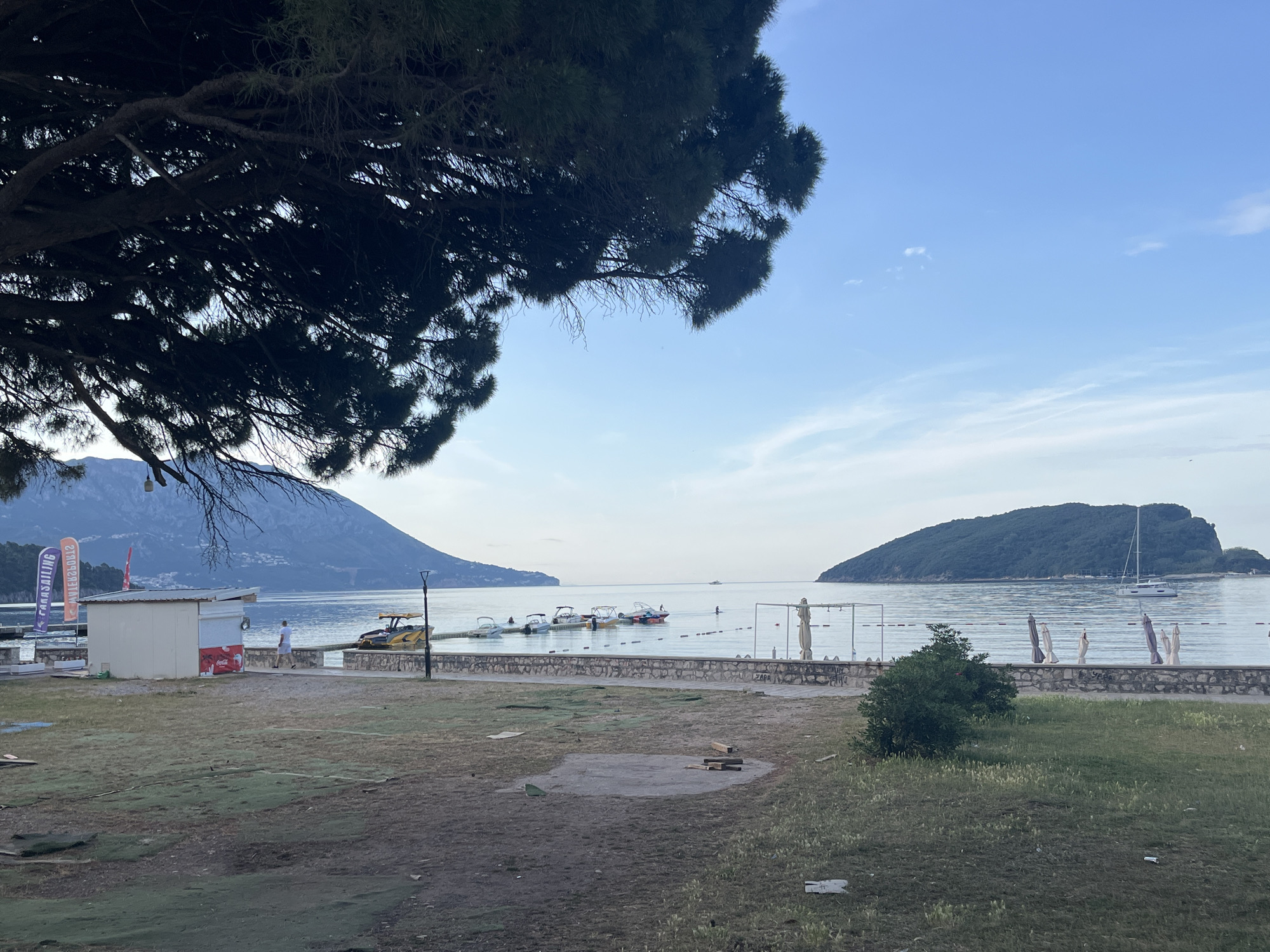 The image captures a view of a beach area in Budva, Montenegro. The scene includes a foreground of grass with patches of bare earth. A tree with dense foliage frames the upper left corner of the image. In the middle ground, there is a paved walkway bordering the beach. A small white building with vending signage is visible. Beyond the walkway, the beach leads to a calm body of water with several boats and a few people swimming. In the background, there's an island and a mountain range visible against a light blue sky. The time of day appears to be morning or early evening, judging by the soft lighting. Beach umbrellas are present along the beach.