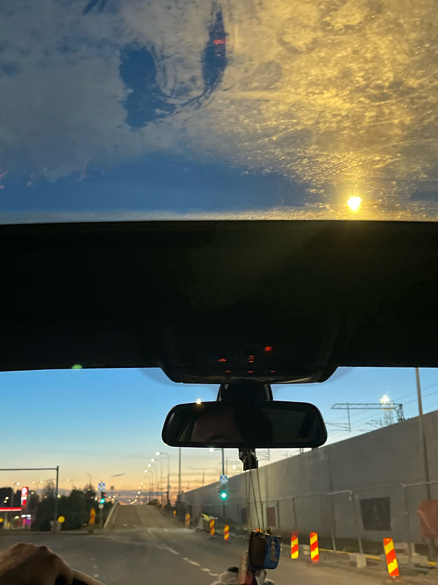 The image is taken from inside a vehicle in Tallinn, Estonia, during what appears to be dusk or early evening. The view is forward-facing, through the windshield and past the rearview mirror. The windshield has some smudges and reflections on it, obscuring parts of the sky and the distant lights.
In the foreground, a portion of the steering wheel and a hand are visible on the left side of the frame. The rearview mirror is mounted in the center of the windshield, and a person's face is partially reflected in it. Above the rearview mirror, the overhead console of the car shows a few illuminated red indicator lights.
Looking through the windshield, a road stretches into the distance, with streetlights lining both sides. The sky is a gradient of blue and orange, indicative of sunset. On the right side of the road, there is a construction site with a tall, gray concrete wall and fencing. Several orange and yellow striped barriers are placed along the side of the road. On the left side of the road, there are some buildings and more streetlights. A traffic light with a green signal is visible in the distance. In the far background, electrical pylons and a large industrial structure are discernible. The overall impression is of a road passing through an urban or suburban area with ongoing development.
