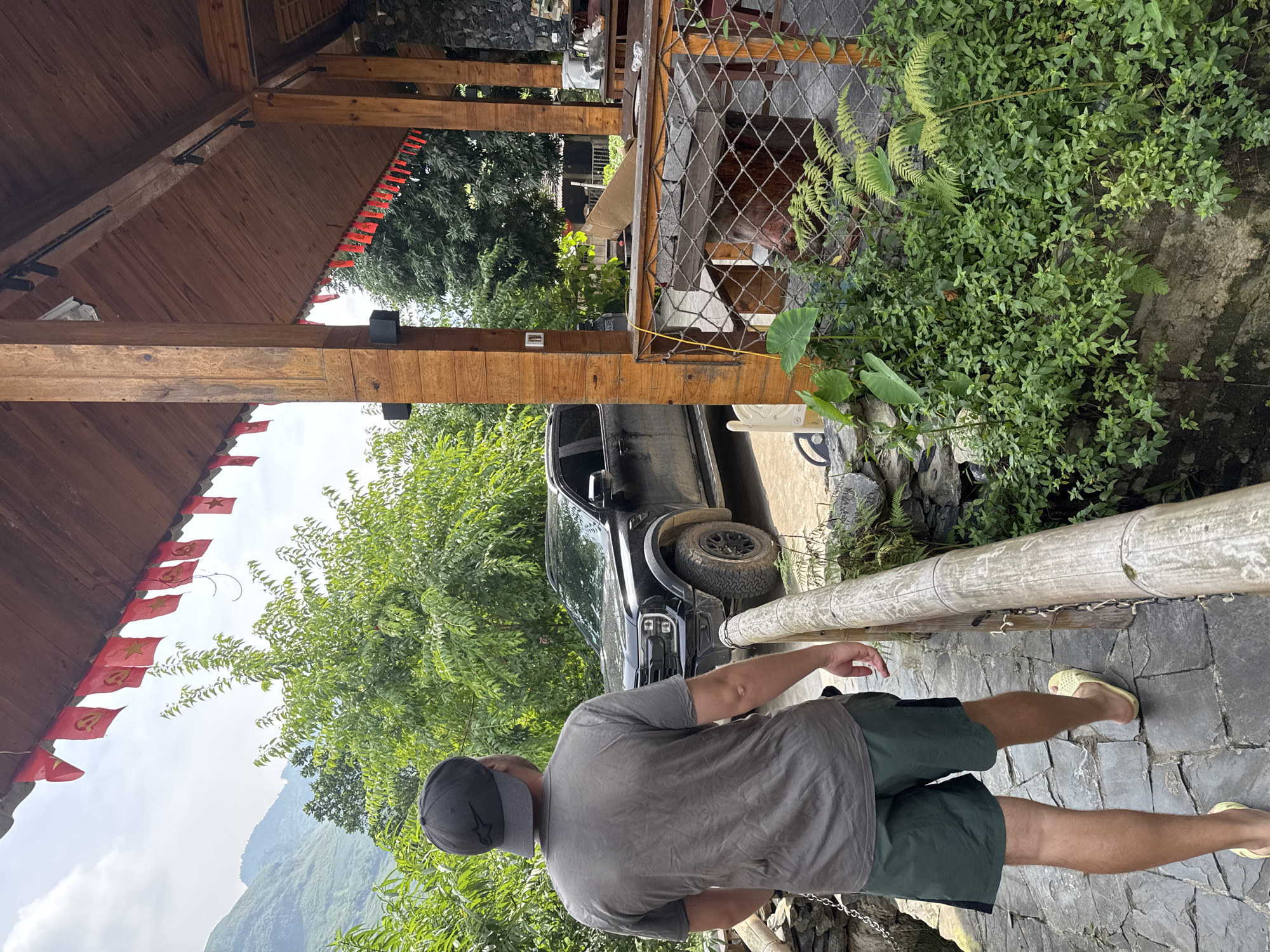 This outdoor image, taken in Du Già Commune, Vietnam, captures a daytime scene with a man walking along a stone path. The man, viewed from behind, wears a grey t-shirt, dark shorts, a dark baseball cap with a small logo, and yellow flip-flops. He is moving away from the viewer, along a path constructed of irregular dark grey stones.
To the right of the path, a wooden railing, appearing to be made of bamboo sections, borders a dense area of lush green vegetation, including ferns and other leafy plants growing over rocks. On the left side of the frame, a sturdy wooden structure with a sloped roof and thick wooden beams suggests an open-air building, such as a patio or restaurant. Hung along the edge of this wooden roof are numerous small red flags; some clearly feature a yellow star, emblematic of the Vietnamese flag, while others display the yellow hammer and sickle symbol.
In the mid-ground, a black pickup truck, possibly an off-road capable model like a Ford Ranger, is partially visible parked amongst green trees. Beyond the immediate foreground and mid-ground elements, large, verdant mountains dominate the distant background under a bright, partly cloudy sky. The overall impression is one of a scenic, natural setting, likely a tourist or guest area, on a clear day. No explicit text is visible, save for the symbols on the flags.