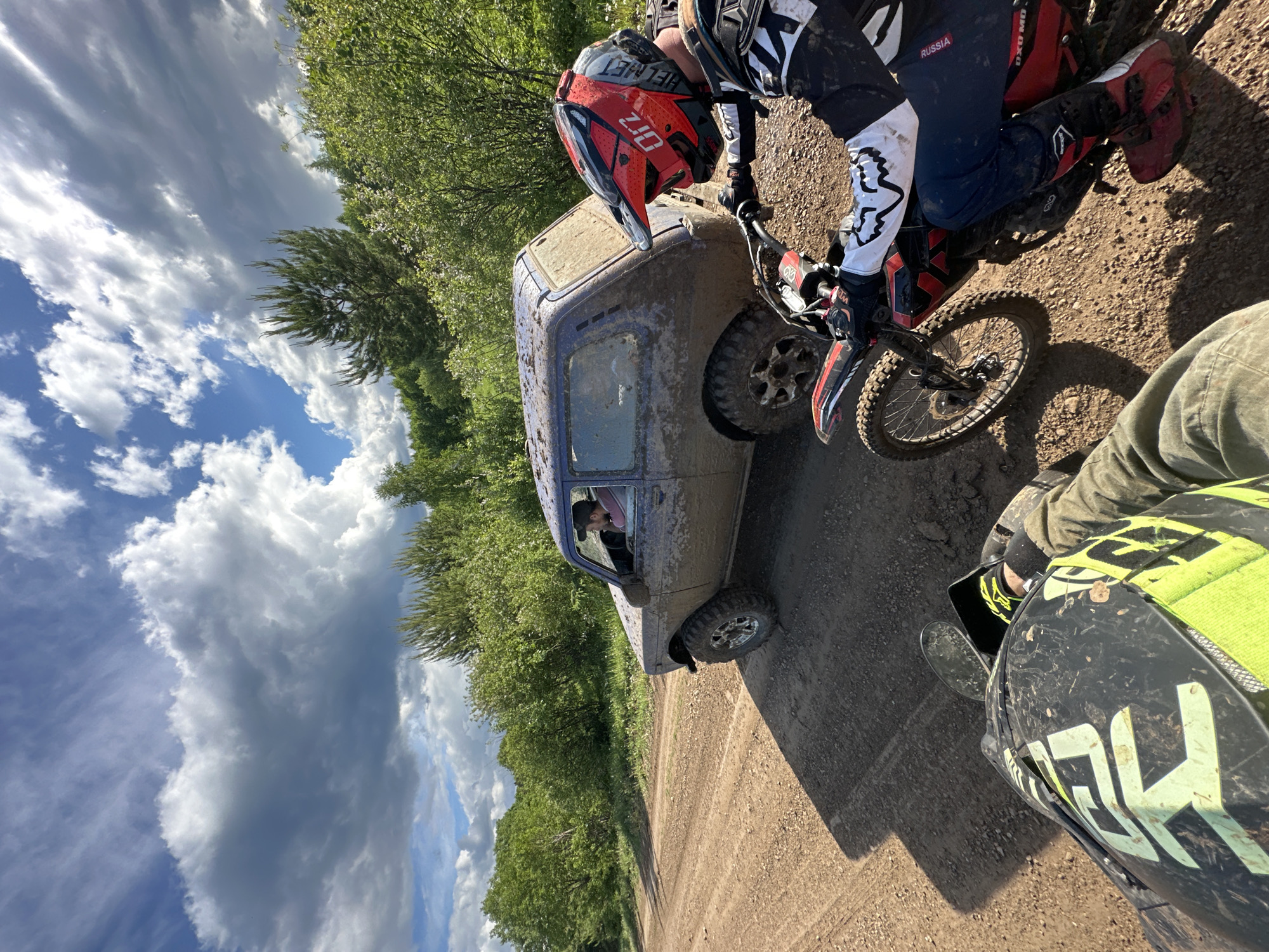 A photograph shows a muddy scene with a dirt road running through a wooded area under a cloudy sky. A blue, mud-covered SUV is parked on the road. A person wearing a red and black helmet and motocross gear is on a red and black dirt bike, positioned next to the SUV. Another person wearing similar motocross gear with a green and black helmet is partially visible in the lower right corner of the frame, suggesting they are also on a dirt bike. The location is Пепеляева, Russia.