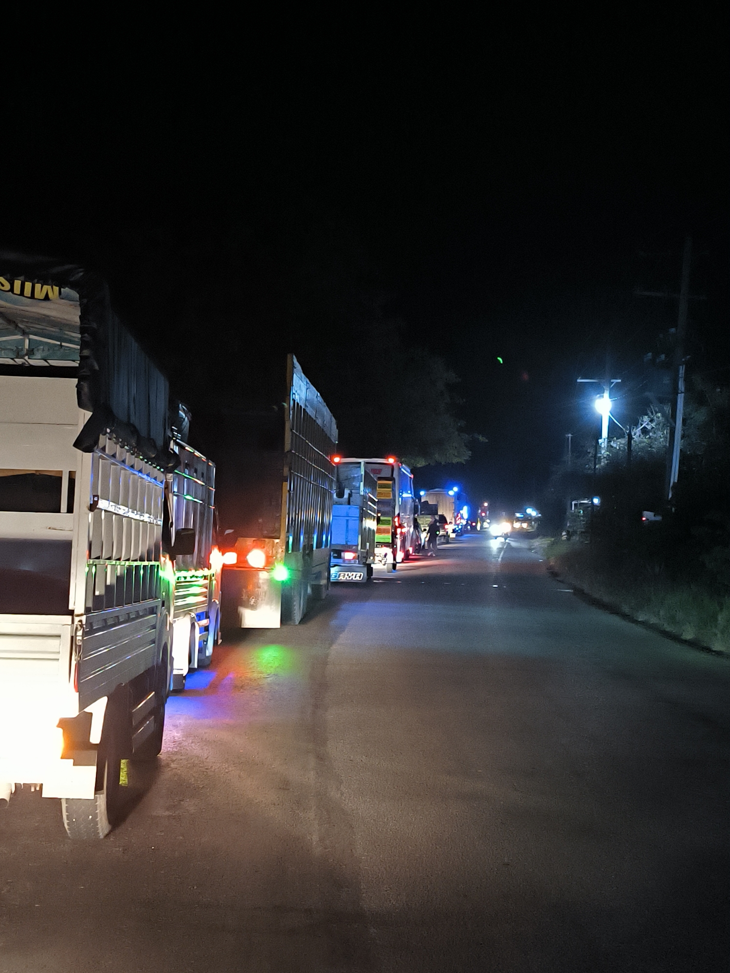 The image depicts a nighttime scene on a paved road in Lhoknga, Indonesia, dominated by a line of large trucks. In the immediate foreground on the left, a white truck features prominently, its side adorned with an array of colorful LED lights in green, blue, and orange, which reflect vibrantly on the dark road surface. A dark tarpaulin cover above its cargo area displays partially visible, inverted text that appears to read "MUSI."
Behind this truck, a series of other large trucks extends down the road, forming what appears to be a queue or a stationary convoy. Many of these trucks exhibit red taillights and various side marker lights. Further down the road, distinct blue lights are visible, possibly indicating emergency services, police, or simply more trucks with custom lighting. One truck in the mid-distance has "JIMU" visible as text on its front.
The setting is dark, with the black night sky suggesting late hours. Illumination comes primarily from the vehicles' lights and a bright street lamp positioned on the right side of the road. This lamp stands beside a utility pole and a dense line of trees and foliage, indicating a rural or semi-urban environment. In the distance, oncoming vehicle headlights are visible, adding to the artificial light sources. The activity suggests either a traffic hold-up, a checkpoint, or trucks parked along the roadside as part of a late-night transport operation. While no clear human figures are discernible in detail, faint shapes suggest people near the distant trucks. The overall scene is calm, with the colorful truck lights providing a striking contrast against the darkness.