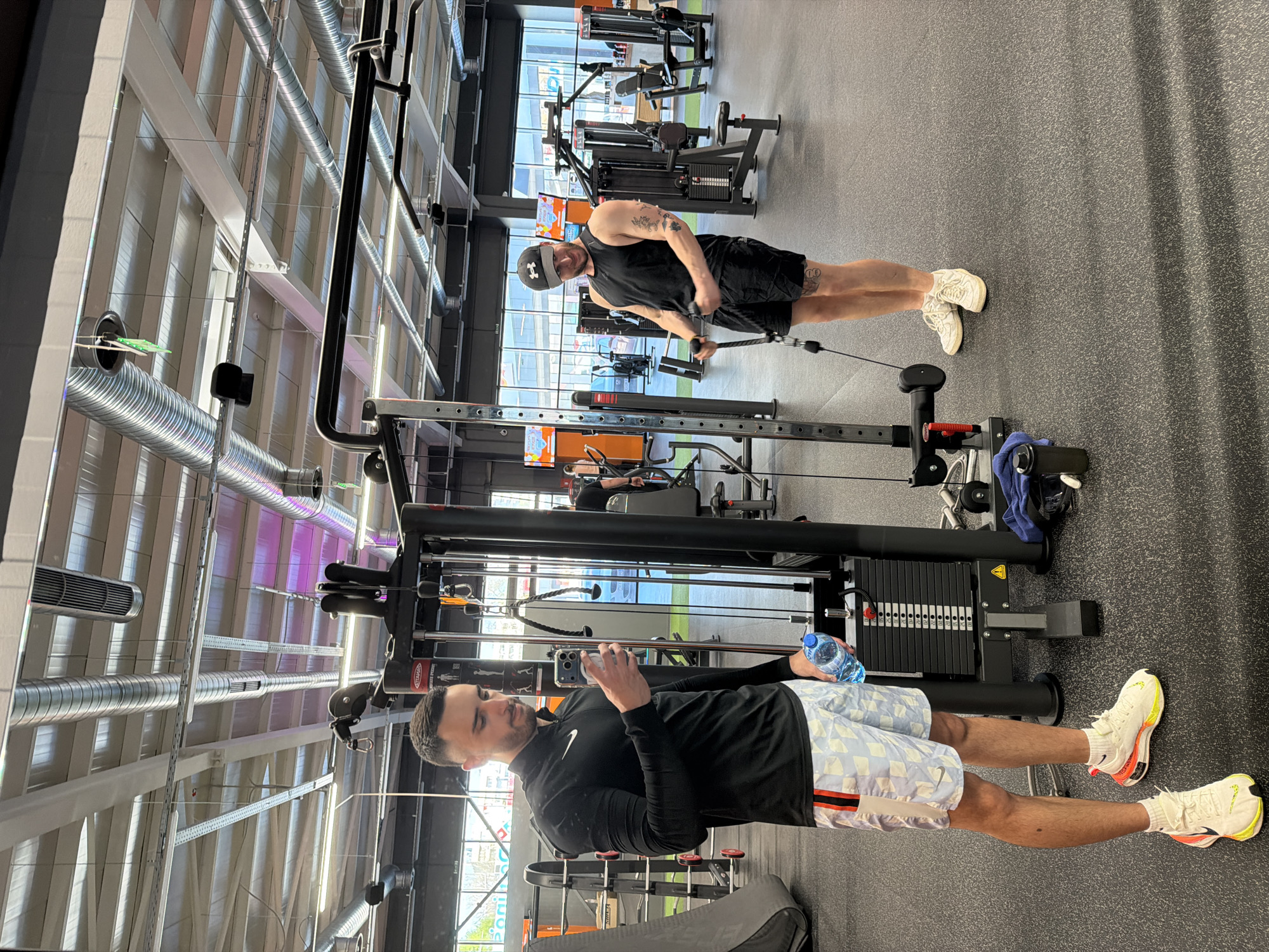 Two men are in a gym. The man in the foreground, on the left, is wearing a black long-sleeved shirt and patterned shorts. He is holding a water bottle and a phone, seemingly taking a picture. The man in the background, on the right, is wearing a black tank top and shorts, with a baseball cap. He is using a cable machine. The gym has a variety of weight machines and equipment. The interior has visible ductwork and fluorescent lighting. The location is inferred to be a gym in Plovdiv, Bulgaria, based on the user request.