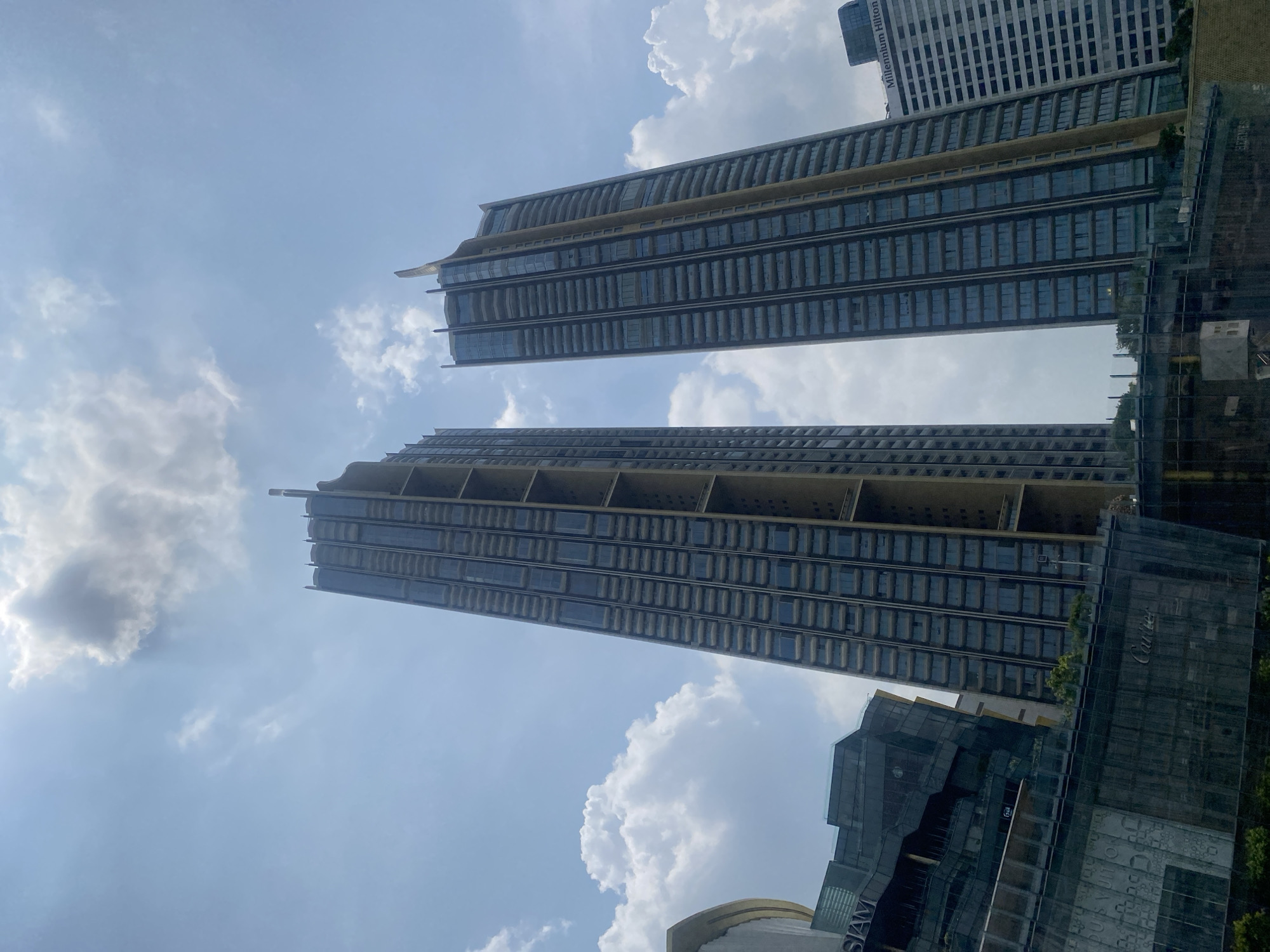 The image shows two tall, modern buildings against a bright blue sky with scattered white clouds. The buildings feature a dark glass facade with horizontal architectural details, giving them a tiered or stacked appearance. They appear to be upscale residential or commercial properties.
Below the buildings on the right side of the frame, there are partially visible modern structures, including a building with "SIAM" visible on its facade and what appears to be a Cartier storefront. These elements, combined with the architectural style of the buildings, strongly indicate that the image was taken in downtown Bangkok, Thailand, most likely in the area around Siam Paragon.