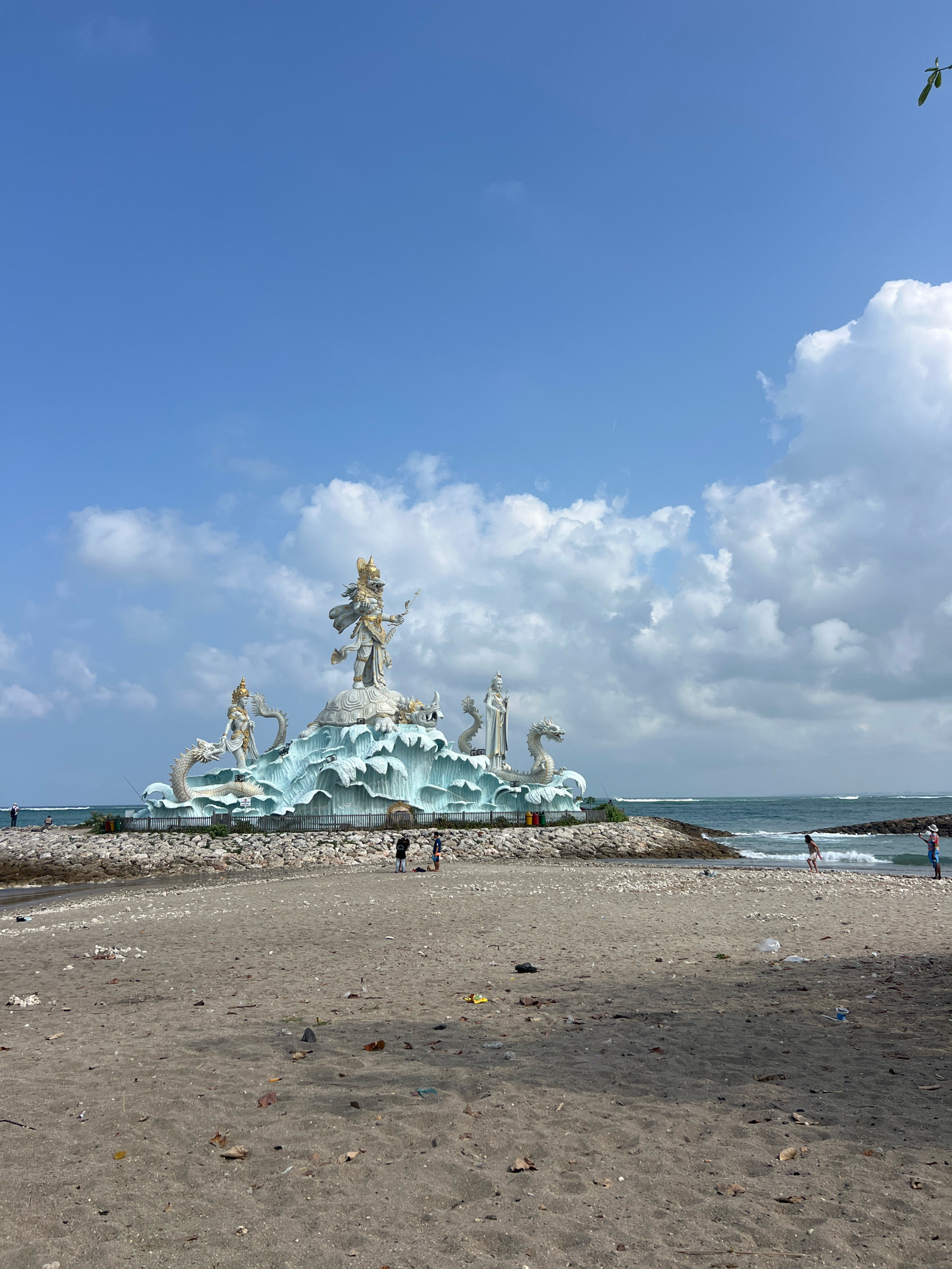 The image shows a statue of a Hindu deity, possibly Dewa Wisnu, on a rocky platform at the edge of the ocean in Kuta, Indonesia. The statue is white and gold, with intricate details, and is surrounded by decorative waves and mythical creatures resembling dragons or serpents. The platform is made of gray rocks and is bordered by a low fence. In the foreground, there is a sandy beach with scattered debris, including leaves and plastic. Several people are visible in the distance, some walking on the beach and others near the water. The sky is blue with scattered white clouds, and the ocean has gentle waves. The overall scene is a blend of cultural monument and natural coastal landscape.