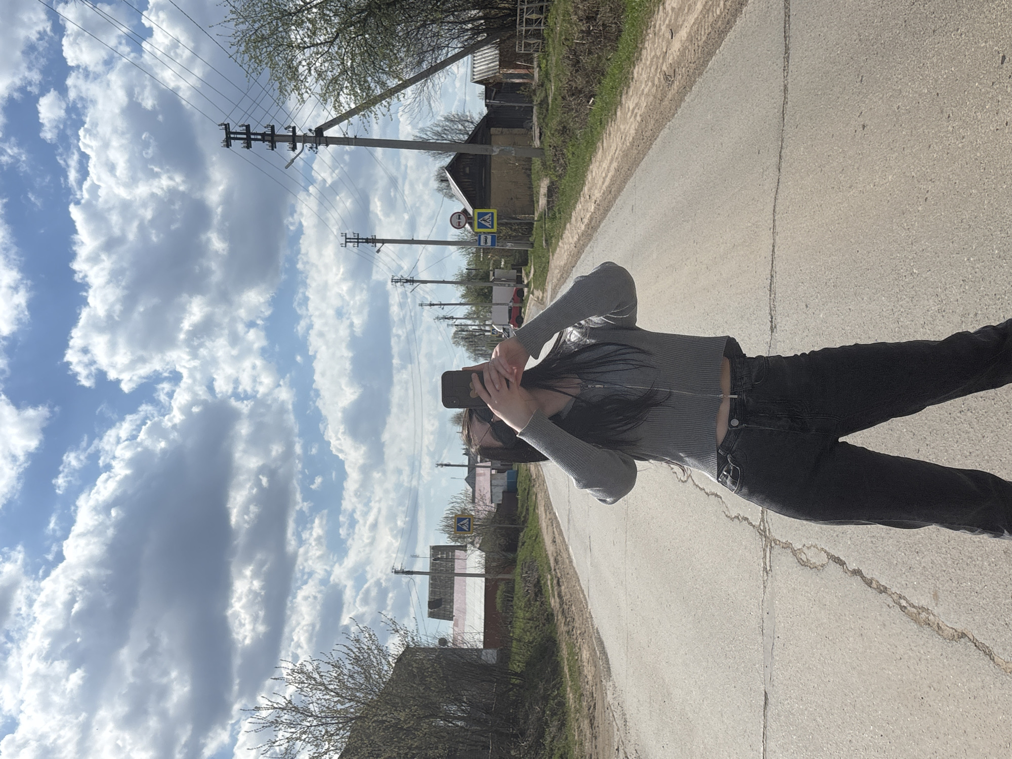 A person stands on a cracked asphalt road in Фоки, Russia, holding a smartphone. The person is wearing a gray top, black pants, and appears to be taking a photo or video. The road is flanked by grassy edges and houses. Overhead power lines are visible against a cloudy sky. A pedestrian crossing sign is present in the background.