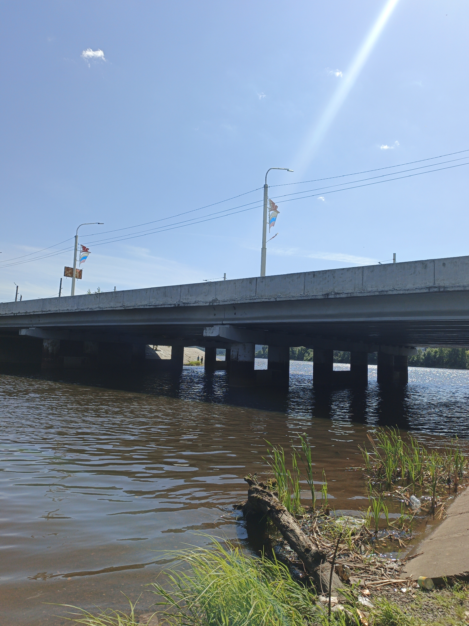The image depicts a wide, concrete bridge spanning a body of dark, murky water. Underneath the bridge, several concrete pillars support the structure, and through the gaps between them, a sunlit riverbank with trees can be seen. There are two small figures visible on the riverbank. On the bridge, two streetlights are mounted on poles, each adorned with a small flag, one appearing to be the Russian tricolor. The sky is a bright, clear blue with a few wispy clouds and a strong sun glare in the upper right quadrant. In the foreground, the water is calm with ripples, and the bank is lined with green reeds and debris, including a large piece of driftwood, some plastic bottles, and other trash. The scene is likely a section of the Ay River in Zlatoust, Russia, as suggested by the context of the request and the typical urban bridge architecture found in Russian cities.