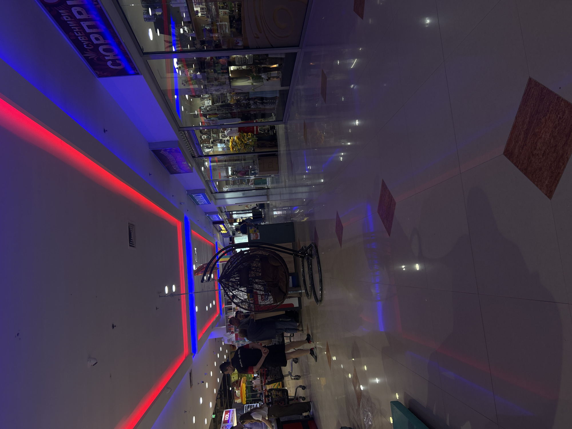 This media file depicts an indoor scene, likely a shopping mall, with a long, brightly lit corridor. The walls are adorned with vibrant red and blue LED lighting, casting a colorful glow on the polished tiled floor. In the foreground, several people are visible, some walking and others gathered near a shopping cart. A distinctive hanging wicker chair with a dark frame is also present in the middle ground, adding a touch of decorative flair to the commercial space.
The overall atmosphere is one of commercial activity and consumerism. The lighting suggests it could be evening or a time when artificial lights are dominant. Reflections on the floor indicate a clean and well-maintained environment.
Visible text includes a sign that appears to read "СПОРТПЛ" or similar, suggesting a sporting goods store or a section related to sports. Another sign in Cyrillic, potentially "КАЧО", is partially visible on a shopping cart. The location context provided indicates Shumen, Bulgaria. There are no indications of the time of day or weather conditions as it is an indoor environment. The emotions of the people are not discernible.
