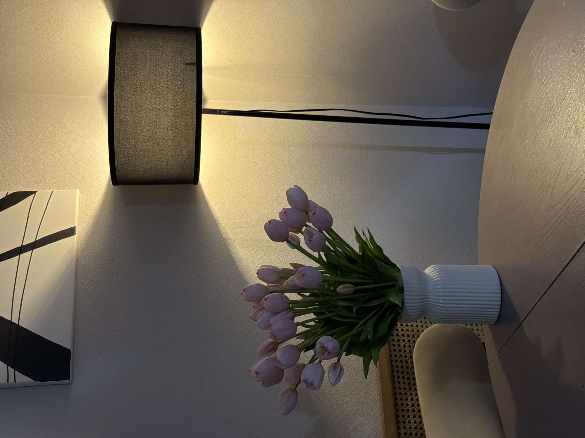 A bouquet of pale pink tulips in a ribbed white vase sits on a round, dusty purple table. Behind the vase, a floor lamp with a black drum shade casts a warm yellow light on the textured white wall. To the upper left, a portion of abstract art with black lines on a white background is visible. The scene is indoors, likely a residential setting in Moscow, Russia.