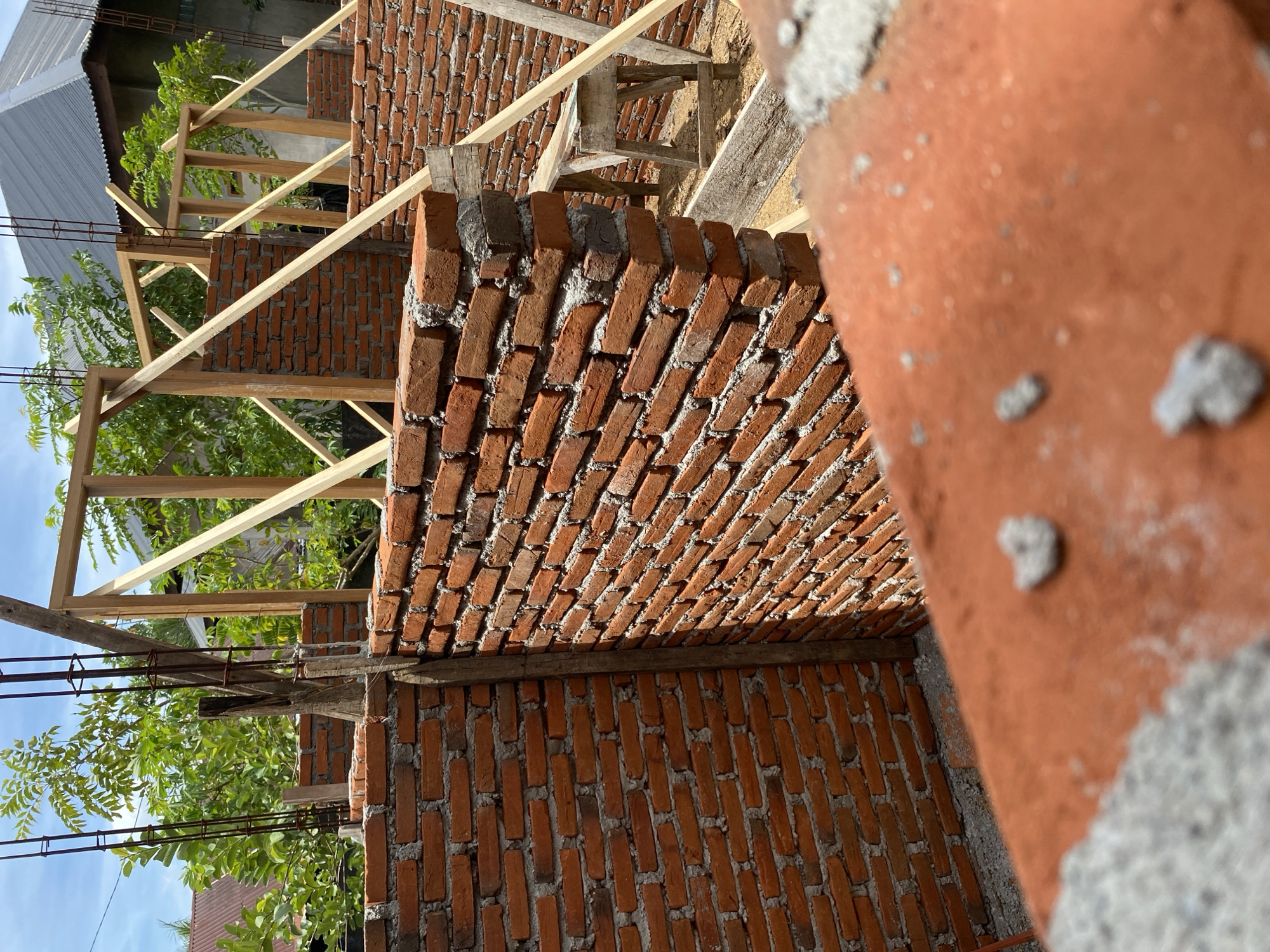 The image captures a construction site in Banda Aceh, Indonesia, focusing on the partial construction of a brick building. The foreground shows a blurred, out-of-focus view of red bricks and gray mortar, suggesting the perspective of someone standing on a newly laid brick surface.
The middle ground is dominated by a brick wall, constructed with red bricks and cement mortar, running diagonally across the frame. To the left, another brick wall is visible, perpendicular to the first. Wooden support beams and metal rebar are integrated into the structure, indicating ongoing construction. Behind these walls, a wooden frame for the building's structure is erected, with diagonal wooden bracing. In the background, lush green trees and a gray, corrugated metal roof of a nearby building are visible, characteristic of the tropical environment of Banda Aceh. Sunlight casts shadows on the brickwork, highlighting the texture and unevenness of the construction. A simple wooden stool or workbench is placed to the right, suggesting tools or materials might have been used there. The overall scene depicts a building in the early stages of construction.