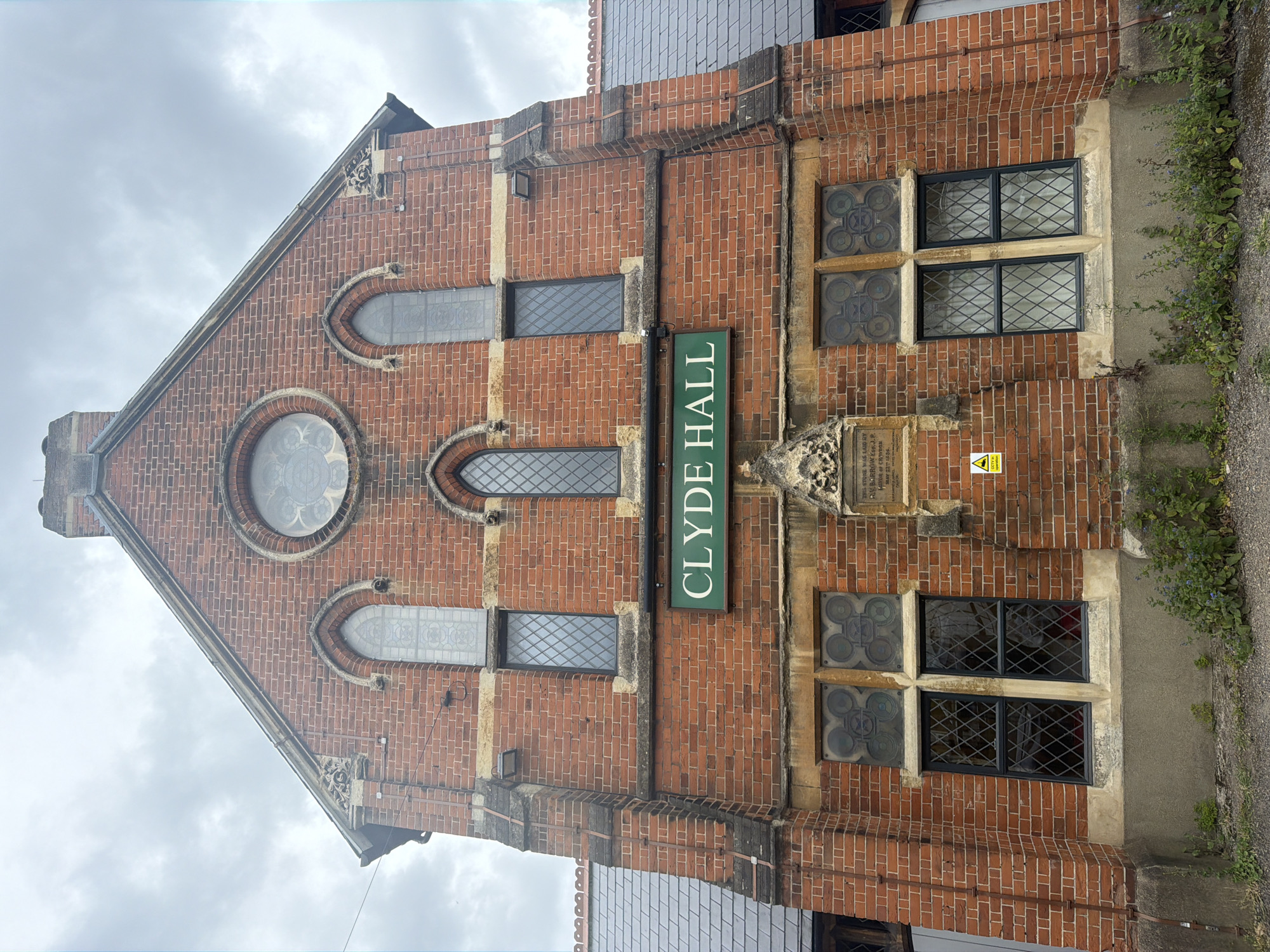 The image captures the exterior facade of Clyde Hall, a red-brick building with distinct architectural features, presumably located in the city of London, United Kingdom. The building is characterized by a symmetrical design, featuring a triangular gable at the top, accented with a circular window and two smaller arched windows. Below these, there are rectangular windows and more elaborate windows to the right of the Clyde Hall sign. A green sign prominently displays the building's name, "CLYDE HALL". A stone plaque is visible, reading, "THIS STONE WAS LAID BY R.V. BARROW ESO. J.P. MAYOR OF CRIVBON MAY 22 1856". A CCTV sign is seen near the plaque. The area surrounding the building features some vegetation. The sky above is overcast.
