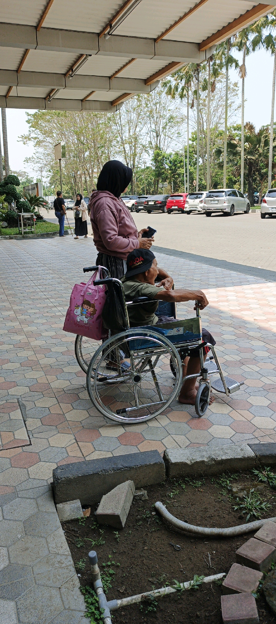 In Ambarketawang, Indonesia, a woman wearing a pink hoodie and black hijab stands behind an elderly man seated in a wheelchair. The woman holds a black smartphone in her right hand, and a pink tote bag with a unicorn illustration hangs from the wheelchair's armrest. The man wears a dark green t-shirt and a black baseball cap with white lettering. They are positioned on a paved walkway with hexagonal tiles in shades of beige, pink, and grey. In the background, cars are parked in a lot, and palm trees and other foliage are visible. The scene appears to be outdoors, under a covered walkway with a corrugated metal roof supported by concrete beams. To the left of the paved area, a patch of dirt contains construction materials and a grey flexible hose.