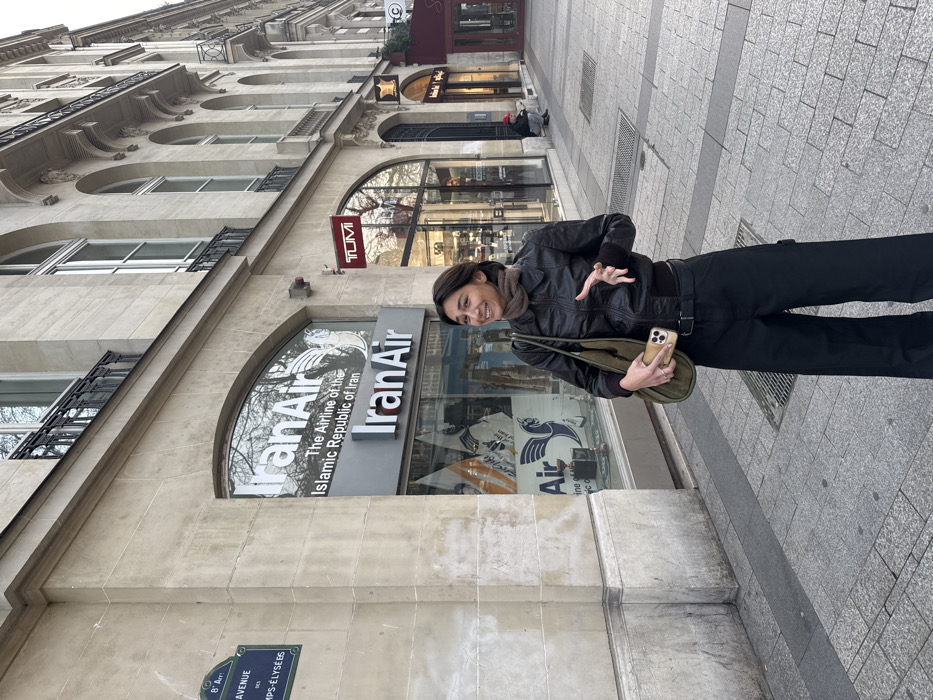 An image captured on the Avenue des Champs-Élysées in the 8th Arrondissement of Paris, France, depicts a female subject standing on a grey tiled sidewalk in front of commercial storefronts. The main subject, a young adult female with dark hair, wears a dark brown leather bomber jacket, a brown knitted scarf, and black wide-leg trousers. She holds an olive-green tote bag over her left shoulder and a light-colored smartphone in her right hand. Her left hand is raised with fingers splayed, mid-gesture, and she is smiling while looking towards her left.
Behind her, a light-colored stone building with ornate Haussmannian architecture features multi-story windows with black iron balconies. The ground floor displays several commercial establishments. Immediately behind the subject is an "IranAir" office, identifiable by large white and red signs reading "IranAir" and "The Airline of the Islamic Republic of Iran," with the company's griffin logo visible in the window displays. Adjacent to IranAir is a TUMI luggage store, marked by a red "TUMI" sign. Further down the street, other storefronts are visible. A blue street sign on the left side of the frame confirms the location as "AVENUE DES CHAMPS-ÉLYSÉES, 8 Arr." In the distance, on the right side of the sidewalk, a person wearing a red hat is seen sitting or crouching near a building entrance. The scene is illuminated by ambient daylight.