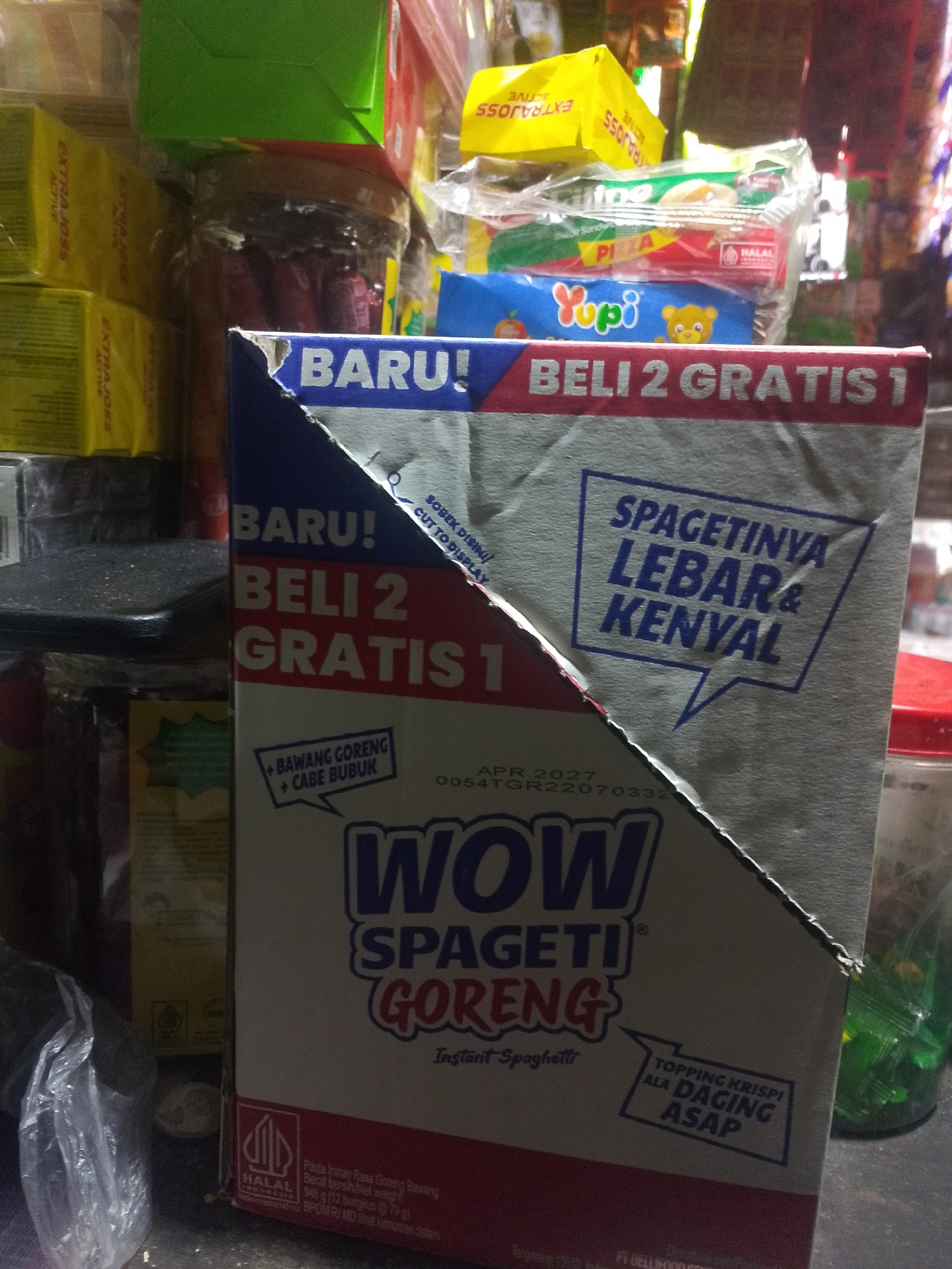 The image shows a retail store shelf with various products, primarily packaged food items, in Palembang, Indonesia. In the foreground, a cardboard box for "WOW SPAGETI GORENG" (fried spaghetti) is prominently displayed. The box is partially opened, revealing the text "BARU! BELI 2 GRATIS 1" (New! Buy 2 Get 1 Free) and "SPAGETINYA LEBAR & KENYAL" (The spaghetti is wide & chewy). Below this, there's a smaller inscription indicating "+ BAWANG GORENG + CABE BUBUK" (+ Fried Onion + Chili Powder) and a date "APR 2027". The box also features a Halal certification logo. Behind the spaghetti box, other products are visible, including stacks of yellow boxes labeled "EXTRA JOSS ACTIVE," a jar containing red items, and various other packaged goods, some with colorful designs and text in Indonesian. The overall environment appears to be a small shop or a market stall, characterized by crowded shelves and artificial lighting.