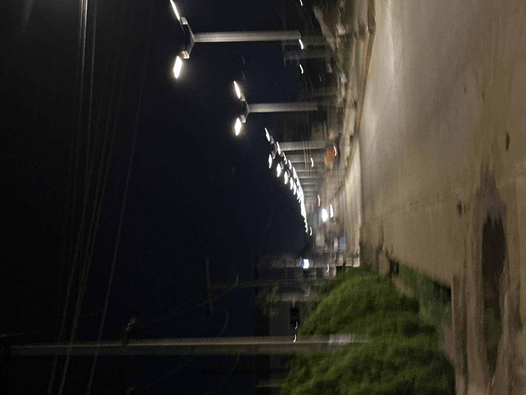 This is an outdoor nighttime scene in Bukuru, Nigeria, showing a street lined with streetlights. The road surface appears concrete with some surface imperfections. Vegetation, likely low-lying shrubs and grass, grows along the roadside. Power lines and utility poles are visible on the left side of the image. The lighting is provided by the streetlights.