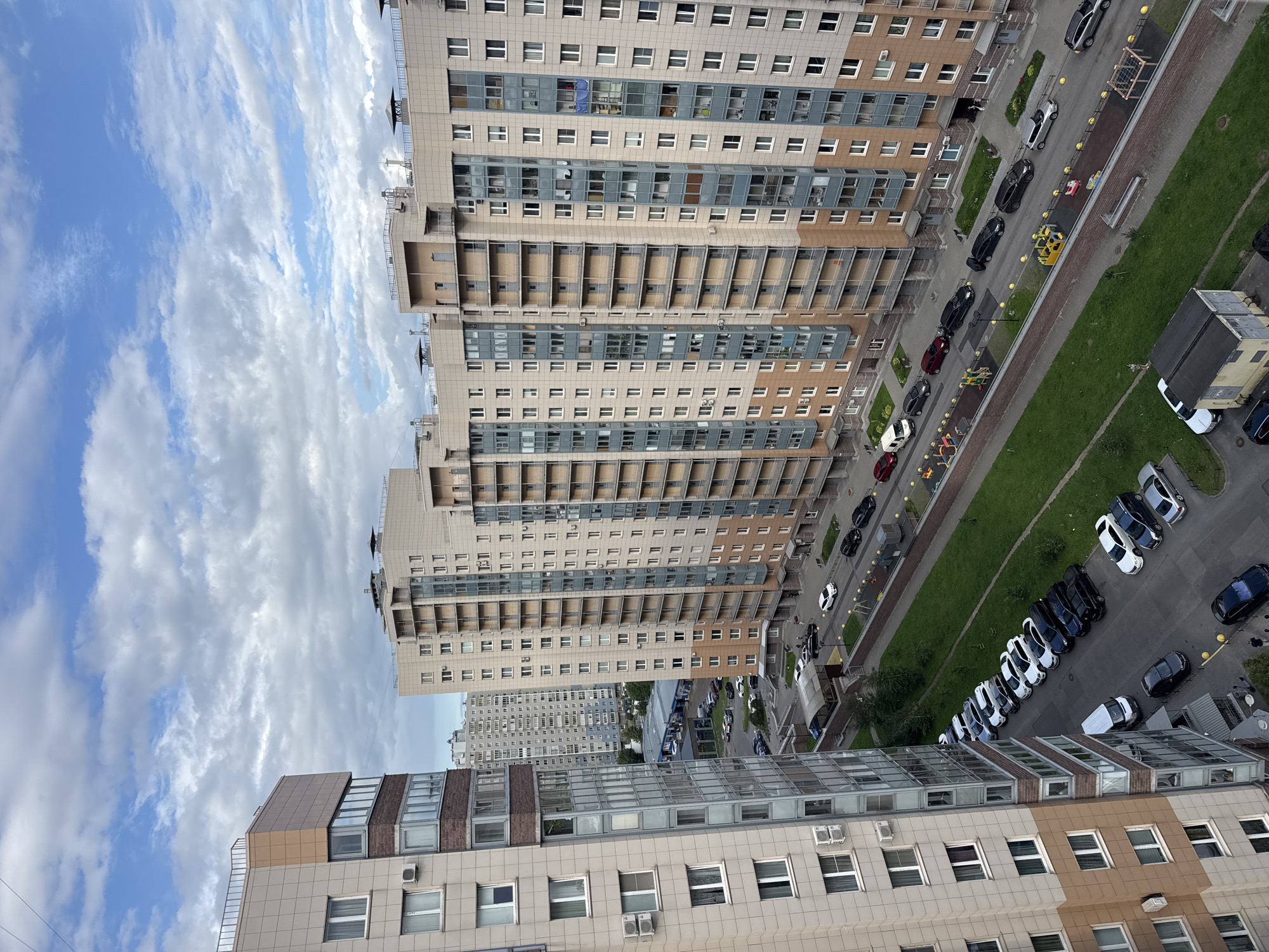 The aerial photograph shows a residential complex with tall apartment buildings in an urban setting under a cloudy sky. In the foreground, a tall, multi-story building with beige and brown paneling and numerous windows is on the left. Adjacent to it, and slightly further back, are several other tall apartment buildings with similar beige facades and balconies. These buildings are lined up along a street with parked cars. The street has several lanes and is bordered by green grassy areas and a paved walkway with children's playgrounds. A variety of cars are parked along the street and in designated parking areas, including sedans, SUVs, and a white van. A few people are visible on the ground, walking on the sidewalks or near the buildings. The environment appears to be a typical urban residential neighborhood with a mix of modern architecture and green spaces.
