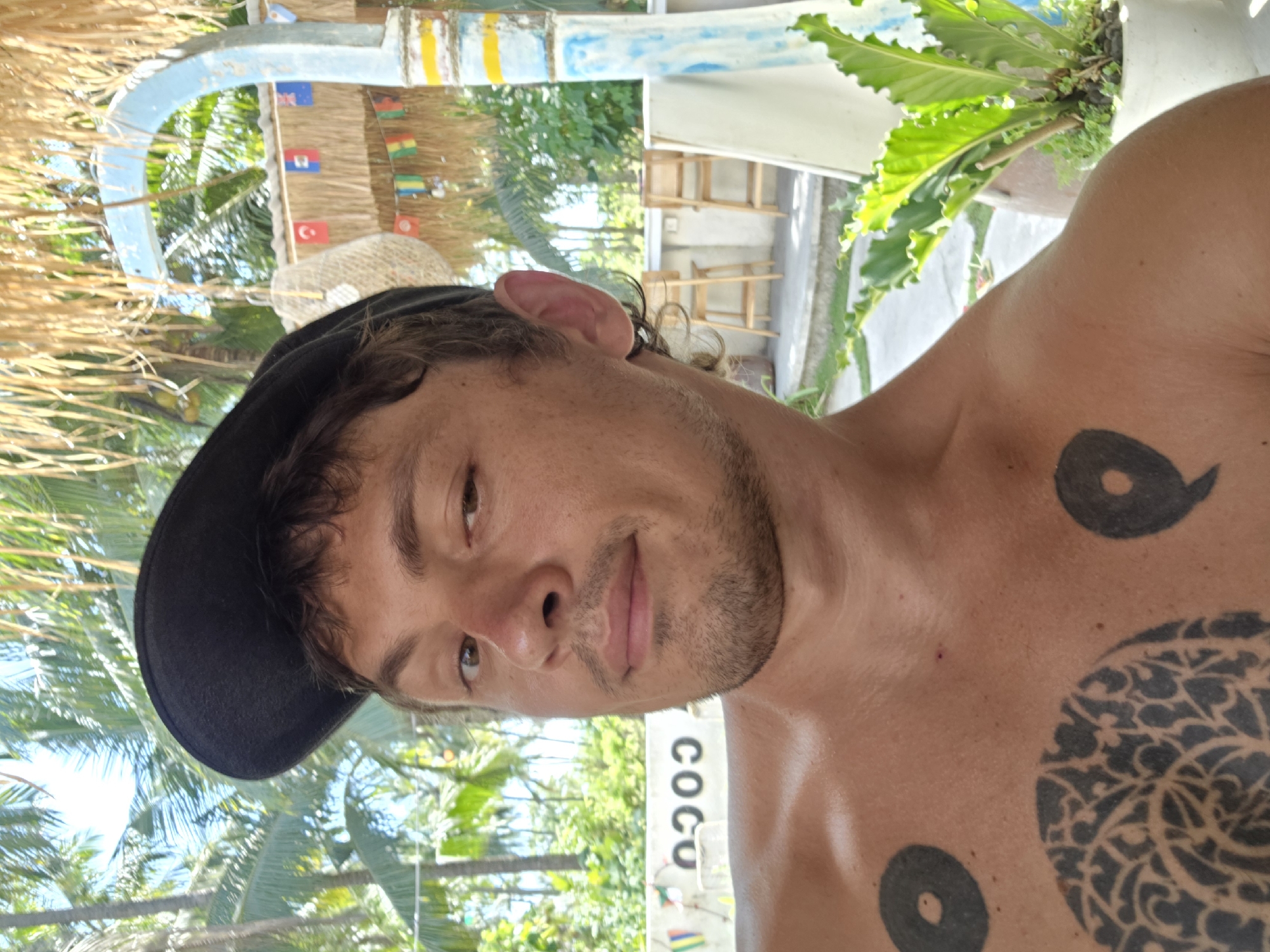 The image shows a shirtless man wearing a black cap, likely in Nusa Penida, Indonesia, as indicated by the "COCO" signage nearby, and tropical vegetation in the background. He has several tattoos on his chest. The setting appears to be outdoors in a tropical location.
