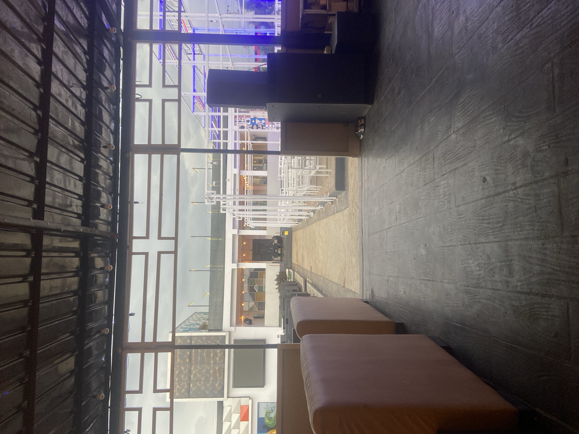 The media file presents an interior or semi-outdoor seating area looking out onto an expansive external space in Gura Top, Nigeria. The scene appears to be a leisure or hospitality venue, possibly a restaurant or event space, currently devoid of people.
In the foreground, a dark, wood-grain textured floor or wall extends into the depth of the image on the right side. On the left, two large, light brown upholstered benches or seating cushions are positioned.
A dominant feature in the midground is a large section of glass windows with intricate geometric, grid-like framing, some of which form a distinct cross pattern. These windows offer a view to the outside. To the immediate left of these windows, a dark, corrugated metal wall or partition is visible, adorned with small, evenly spaced circular lights. Large, dark audio equipment or speakers are also present near the window section.
Beyond the windows, an outdoor environment unfolds. A long, light-colored pathway or paved area leads into the distance, bordered on both sides by numerous white outdoor tables and chairs, set up in rows. Further in the background, modern buildings with varied architectural details and textured facades are visible. Slender lampposts are interspersed throughout the outdoor area. The sky, viewed through the windows, appears overcast, contributing to a soft, diffused light over the scene. There is indistinct, undecipherable text on a distant blue sign.