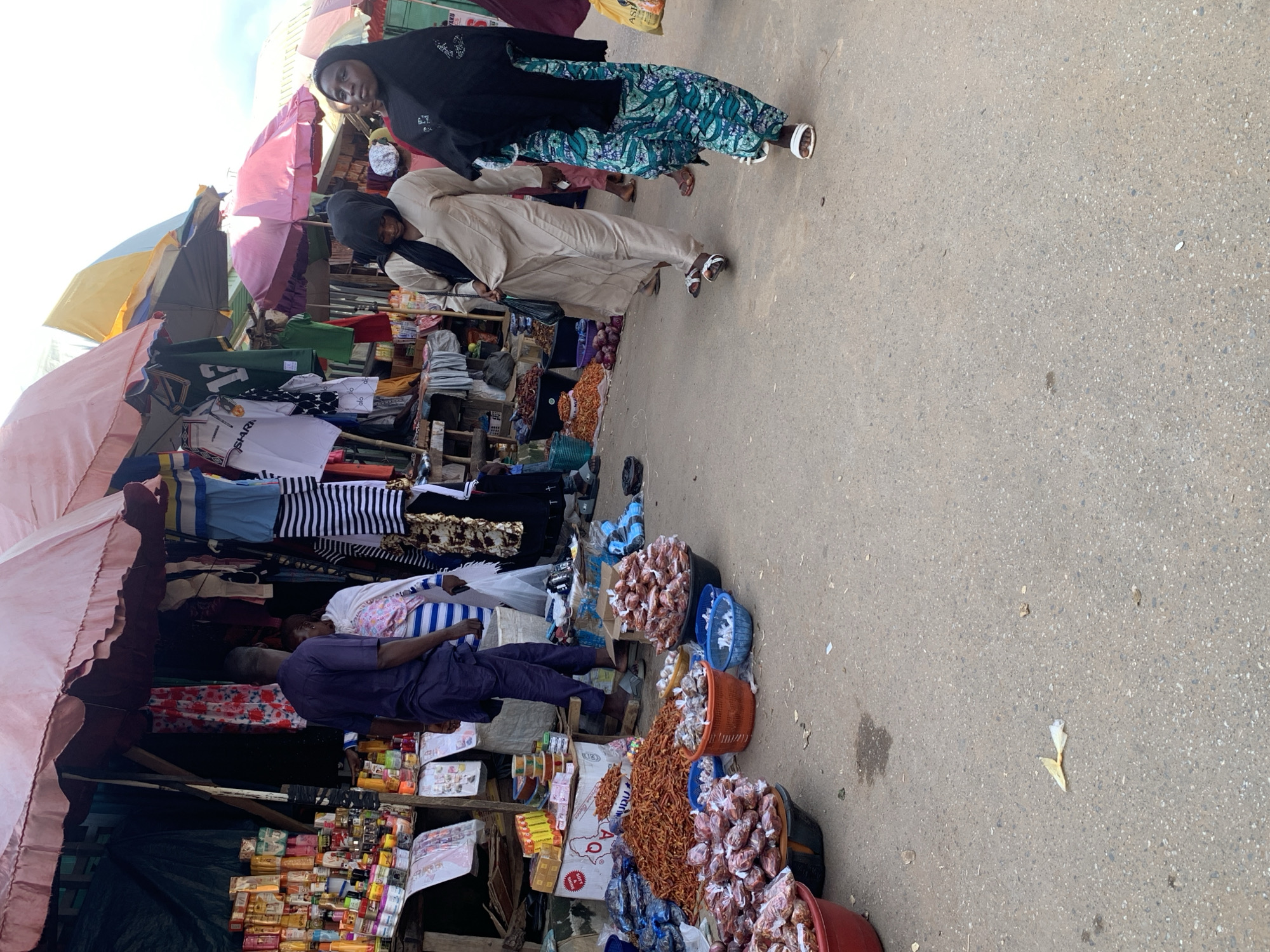 The image depicts a bustling open-air market scene in Akwanga, Nigeria. In the foreground, several large red and black plastic basins are filled with dried red chili peppers, some of which are also bagged. A smaller orange basket, also containing dried chili peppers, sits between two blue plastic baskets, one empty and one with what appear to be bagged spices. Behind these, a substantial pile of dried red chili peppers is visible on the ground, next to a large white cardboard box with red lettering.
The market stalls are characterized by their awnings made of fabric in various colors, including pink, yellow, and red. Merchandise is displayed on wooden shelves and hanging from structures. On the left, a stall is laden with a variety of packaged goods, including colorful boxes and bottles that appear to be toiletries or food items. Further back, clothing items like striped t-shirts and other garments are displayed.
Several individuals are present in the scene. On the left, a man in a dark blue tunic and trousers is standing near a stall, facing away from the camera. Beside him, a person wearing a white and pink striped garment and a white head covering is also visible. Further into the market, a woman in a beige hijab and dress is walking, holding a black plastic bag. Another woman to her right is wearing a black hijab and a green and teal patterned dress, also walking. The ground of the market is paved with asphalt, showing some stains and scattered debris, including a crumpled yellow piece of paper. The overall impression is one of a lively marketplace with vendors selling agricultural produce and other goods.
