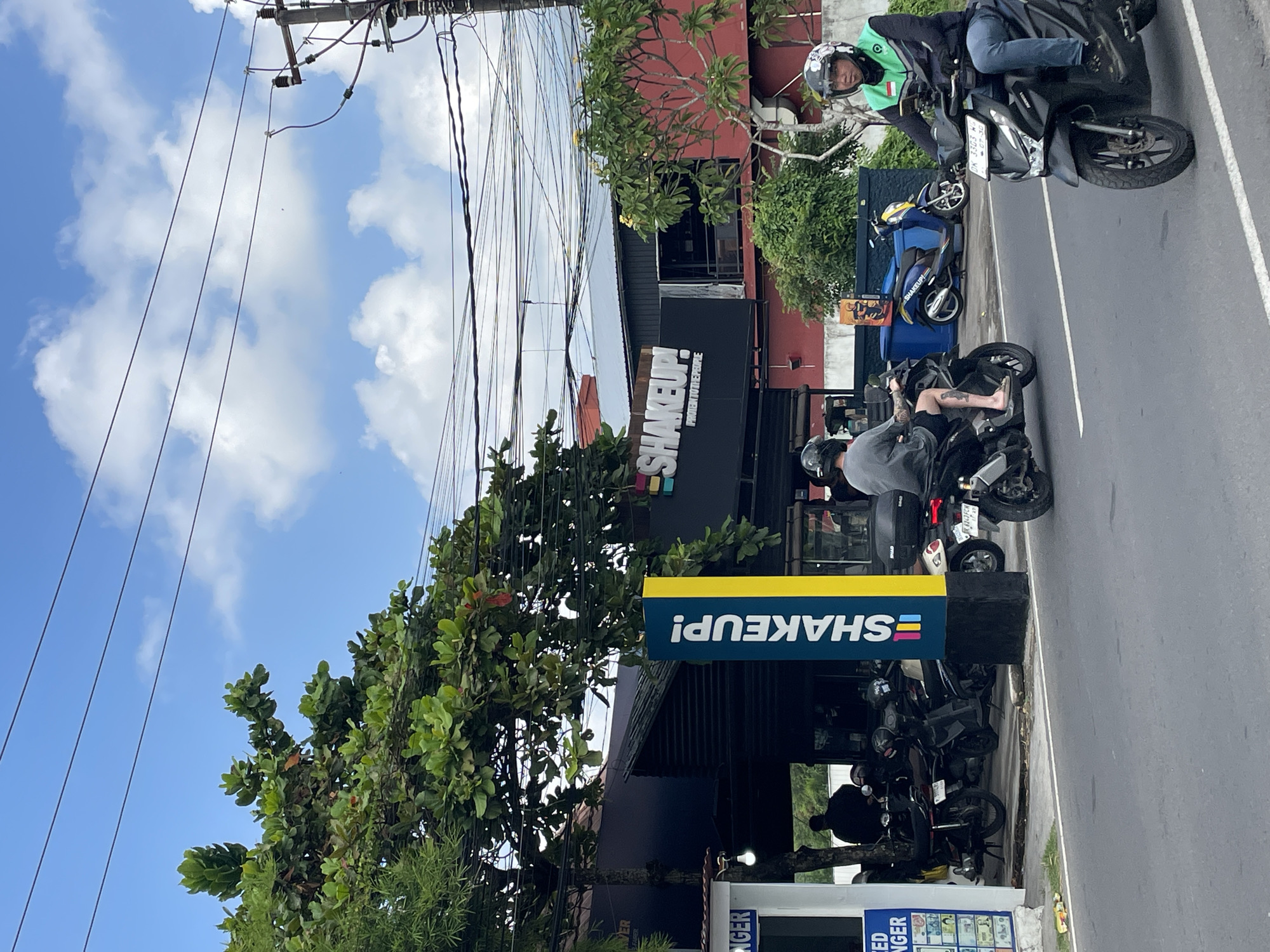 In Mengwi, Indonesia, a street scene unfolds under a blue sky dotted with white clouds. Power lines crisscross overhead, supported by a utility pole on the right. A building with a dark facade and a red upper story displays a prominent sign that reads "SHAKEUP!" with the tagline "PROTEIN TO THE PEOPLE". In front of this building, a vertical sign with a rainbow stripe and the words "SHAKEUP!" stands out.
Several parked motorcycles are visible in front of the building, suggesting a local hub. On the street, two individuals are riding motorcycles. In the foreground, a person wearing a helmet and a green and black vest rides a black motorcycle. Further back on the left, another individual, also wearing a helmet, rides a black motorcycle. A blue scooter with the "SHAKEUP!" logo is parked to the left of the red building. Trees with green foliage line the area. A small white structure with signage is visible on the far left. The road is paved with white lane markings.