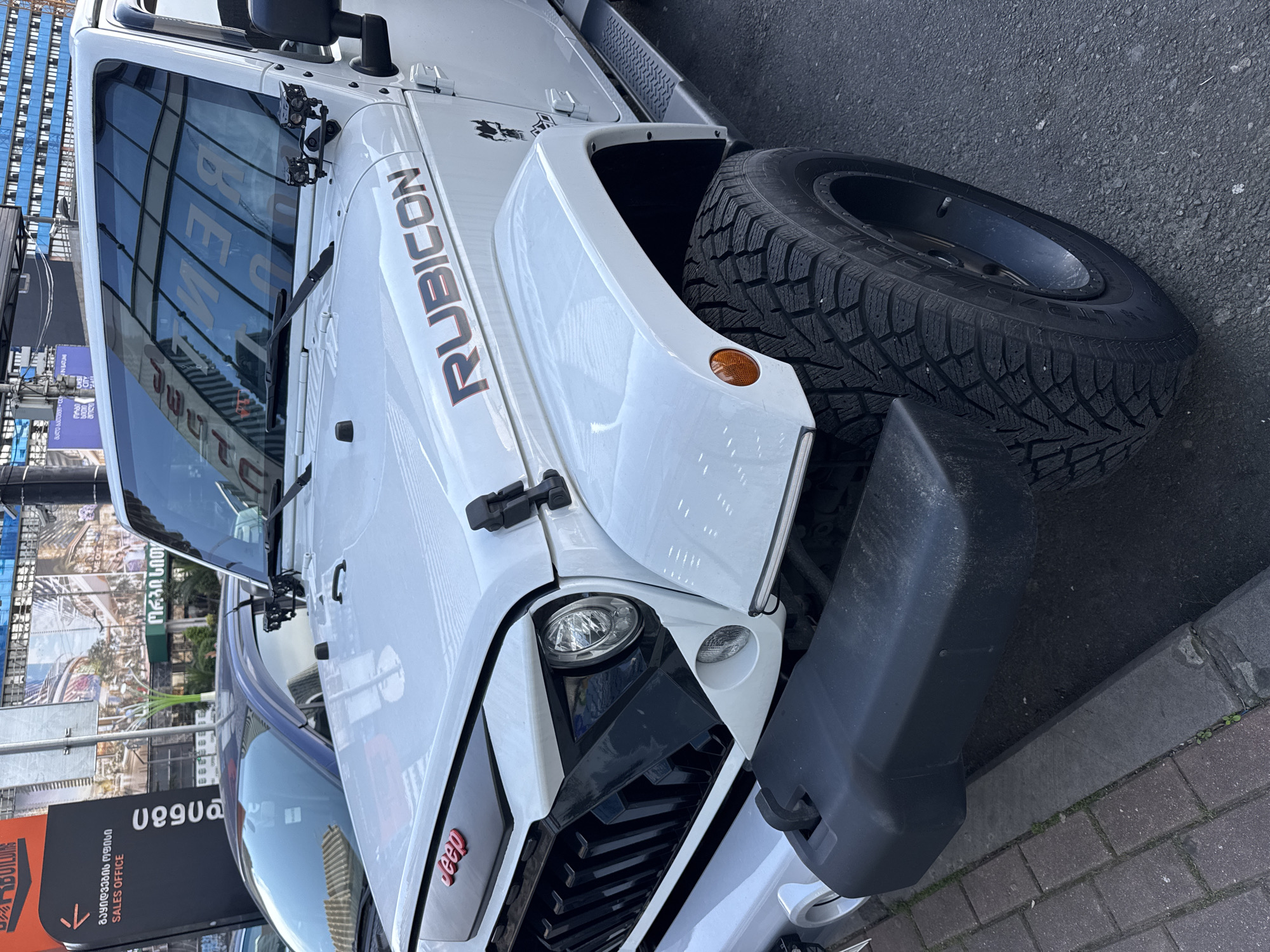 A close-up, medium-angle shot features a white Jeep Rubicon parked on a brick sidewalk. The front right tire is visible, along with the front grill and headlight. A black bumper is present. A building with a glass facade is reflected in the windshield. A sign written in Georgian, which includes “საცხოვრებელი კომპლექსი გაყიდვების ოფისი SALES OFFICE,” is partly visible in the background, placing the location in Batumi, Georgia.