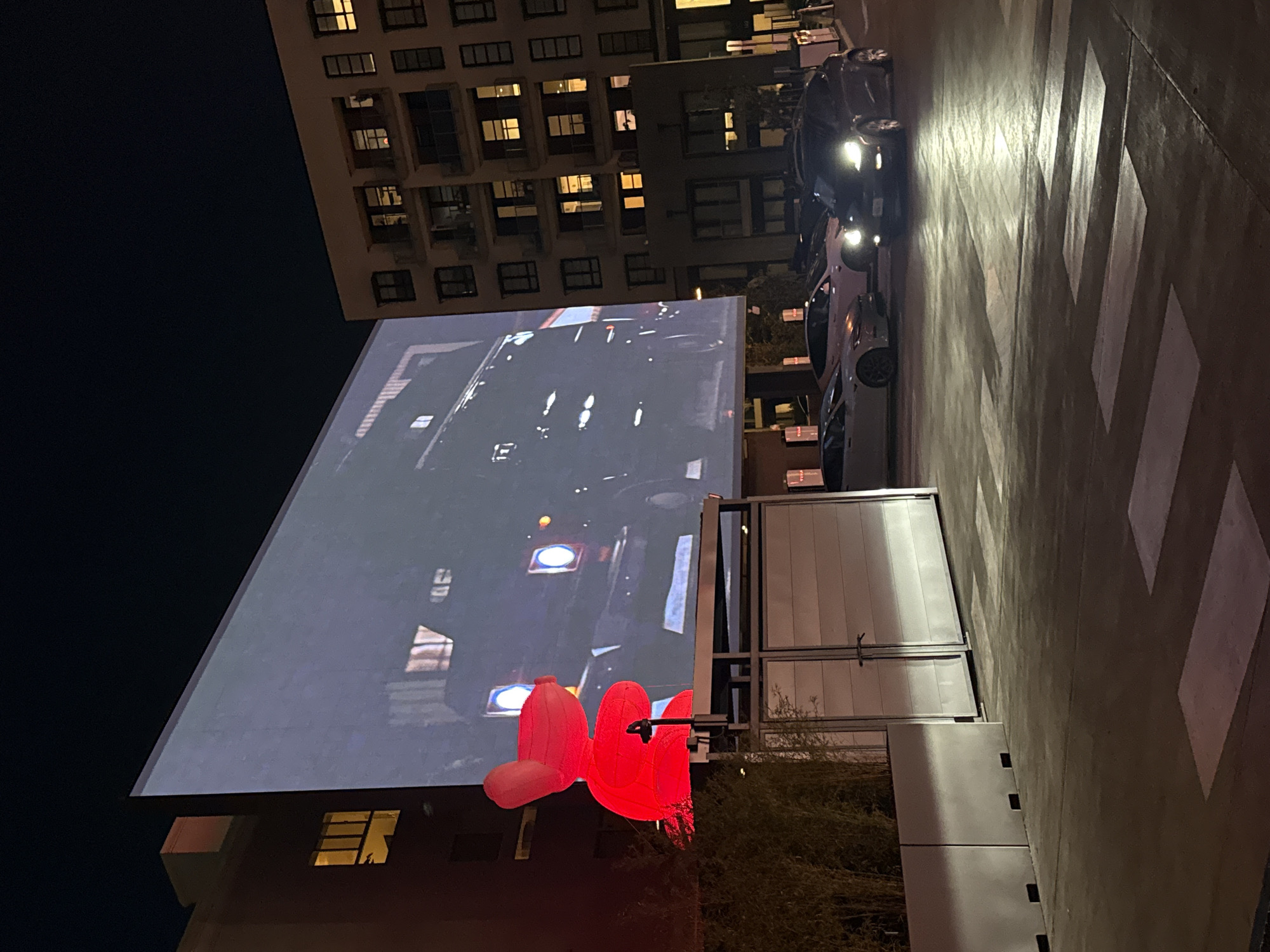 The image depicts a nighttime street scene in West Hollywood, California, United States. A large screen displays a moving car video, presumably an advertisement. In front of the screen is a steel-framed structure with reflective white panels. A red sculpture of lips sits near the structure's base. A building with multiple illuminated windows stands in the background. Several cars are parked on the street, and a crosswalk is visible on the pavement.