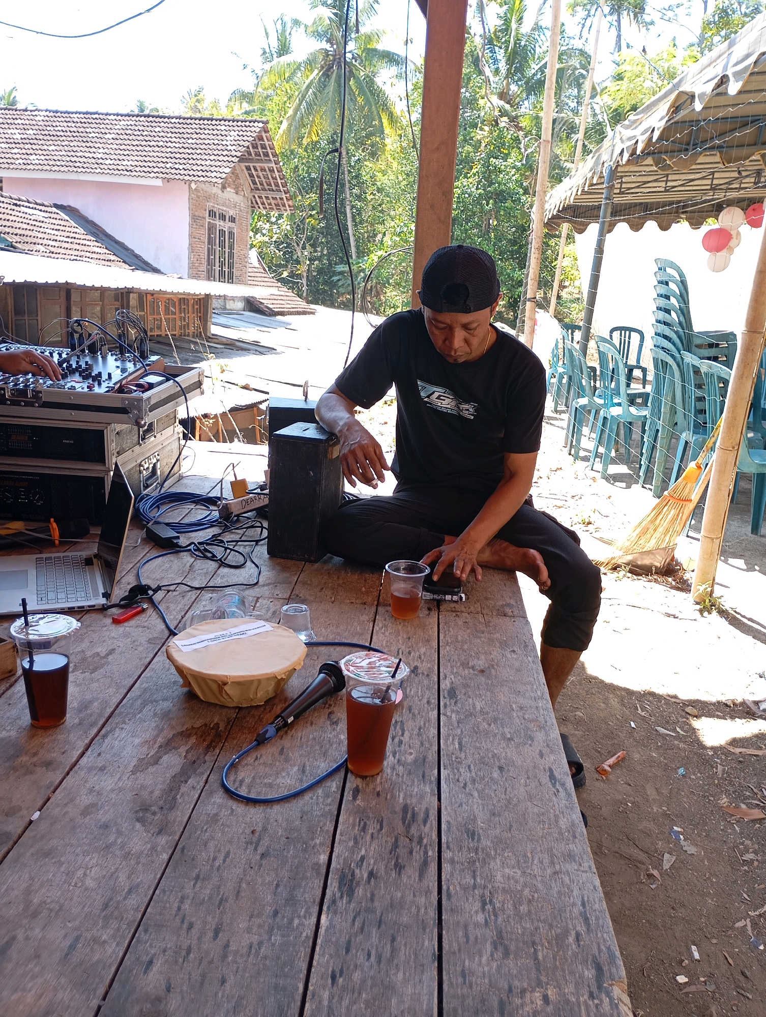 A man in a black t-shirt and black cap sits on a wooden table in an outdoor setting, looking down at a cigarette pack he is holding. In front of him are two plastic cups of iced tea, a microphone, a speaker, and a laptop. A sound system is set up to his left, with cables running across the table. In the background, there are stacked chairs under a tent, palm trees, and a building with a tiled roof, suggesting a location in Ketepung, Indonesia. The scene appears to be a casual gathering or event.