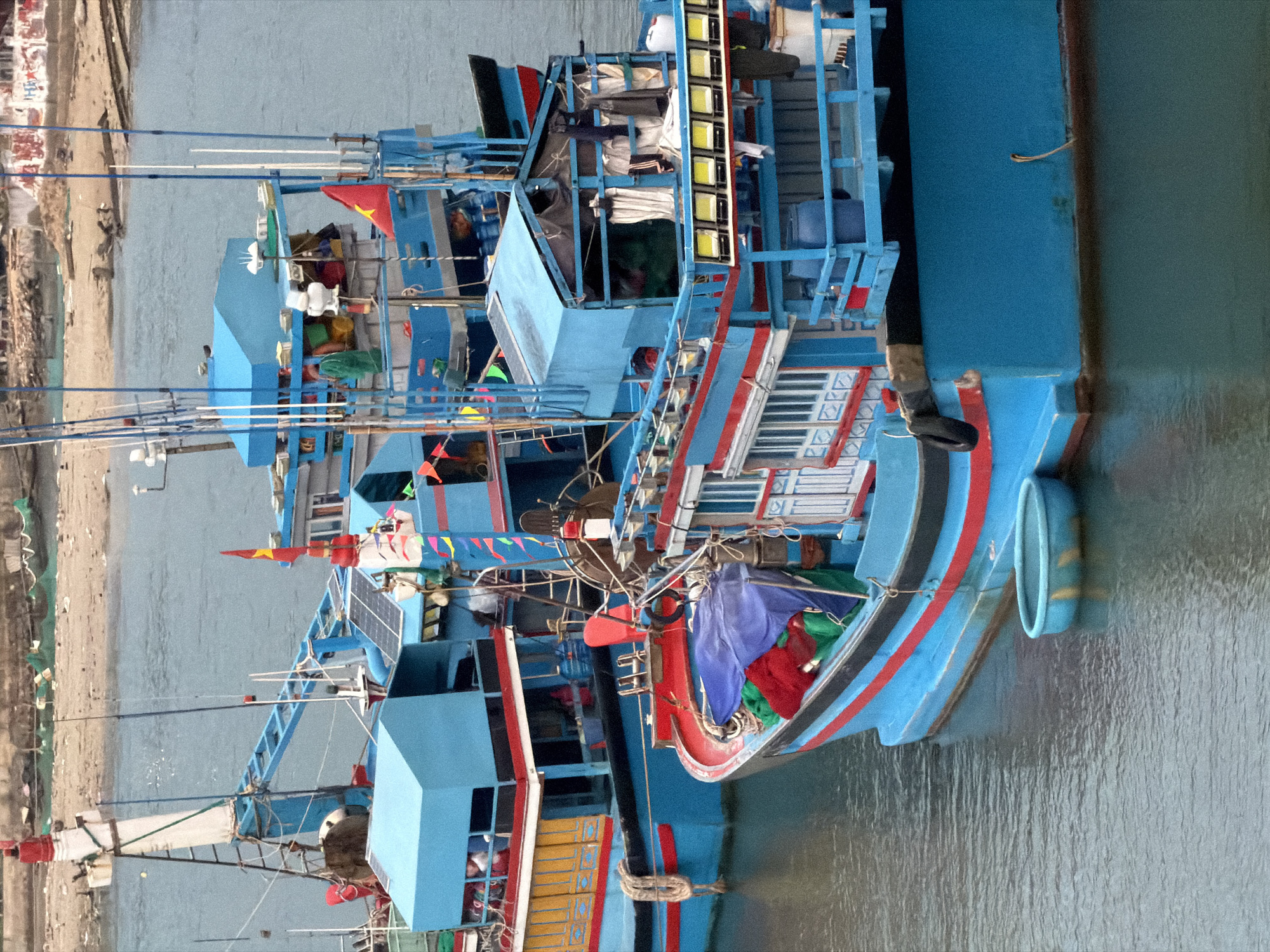 This media file captures a vibrant scene of several fishing boats moored in calm water, likely in a harbor or river in Ninh Chu Ward, Vietnam. The image is oriented with the boats facing right, and the horizon line is vertical on the right side of the frame.
The boats are the primary subjects, painted predominantly in a striking bright blue. They feature multi-tiered superstructures, including cabins and decks, adorned with red, black, and white accents. The main boat in the foreground displays intricate white decorative patterns along its side. Each vessel is heavily equipped with numerous tall masts, fishing poles, and various lines.
Notable details include:
* **Flags and Bunting**: Several Vietnamese flags (red with a yellow star) are visible flying from the boats. Colorful triangular bunting is strung along railings, adding a festive touch.
* **Solar Panels**: At least one boat is equipped with a solar panel array mounted on its upper deck, suggesting modern power solutions.
* **Onboard Items**: Bundles of materials, possibly nets or tarpaulins, in blue, green, and red colors, are stowed on the decks. Lights, possibly for night fishing, are also visible on one of the larger structures.
* **Fenders**: Black rubber tire fenders and large light blue ring-shaped fenders are seen hanging from the boats' sides, protecting them from impact.
The boats are floating in calm, slightly murky blue-green water, which gently reflects their colors. In the background, a sandy or muddy shoreline is visible, with indistinct structures or piles of debris further back. The lighting suggests it is daytime with bright, clear weather. No people are distinctly visible, and no specific activity beyond being moored is clearly taking place. No discernible text is visible on the boats or in the background.