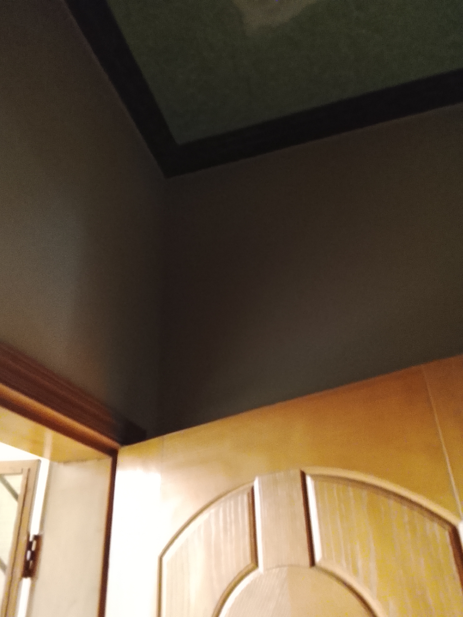 The image shows the corner of a room with a wooden door frame and door. The ceiling is dark, with a recessed section with a darker, framed panel. The lower portion of the frame of a window is visible to the left. The lighting is dim, giving the surfaces a warm tone.