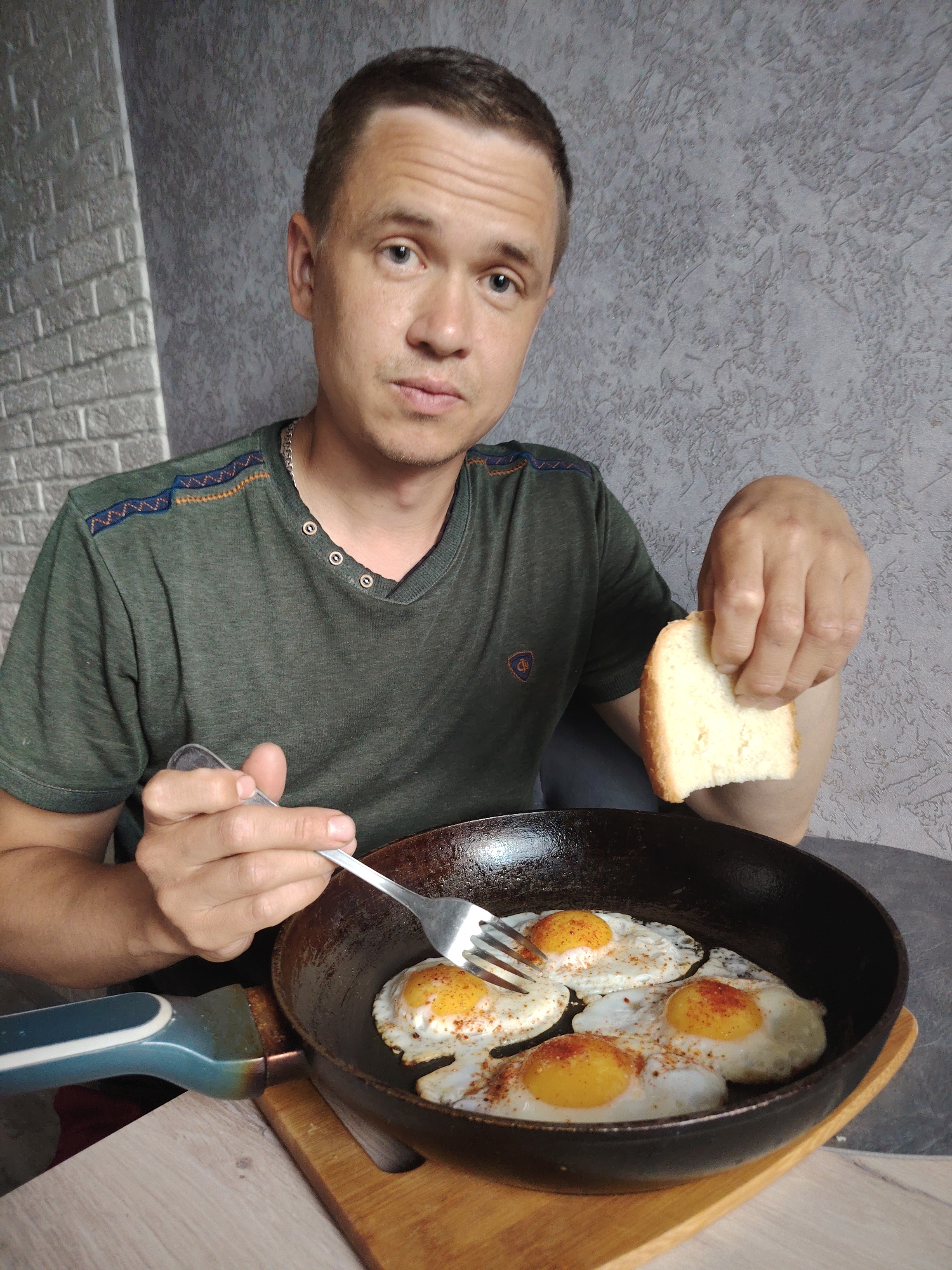 A man with short hair and a neutral, direct gaze is captured in an indoor setting, seemingly about to eat. He wears a dark green short-sleeved t-shirt with decorative patterns on the shoulders and a small, indistinct emblem on the left chest. A silver chain is visible around his neck.
In his left hand, he holds a silver fork poised over a black frying pan that contains four sunny-side-up fried eggs. The eggs have bright orange yolks and white albumen, and appear seasoned with a reddish spice. In his right hand, he holds a slice of white bread. The frying pan rests on a wooden cutting board or trivet, placed on a light-colored wooden table.
The background features two distinct wall textures: a light gray brick pattern on the left, and a darker, textured gray surface on the right, suggesting an interior space like a kitchen or dining area. The man's pose and the meal indicate an everyday activity, likely breakfast or brunch. No visible text is discernible in the image.
