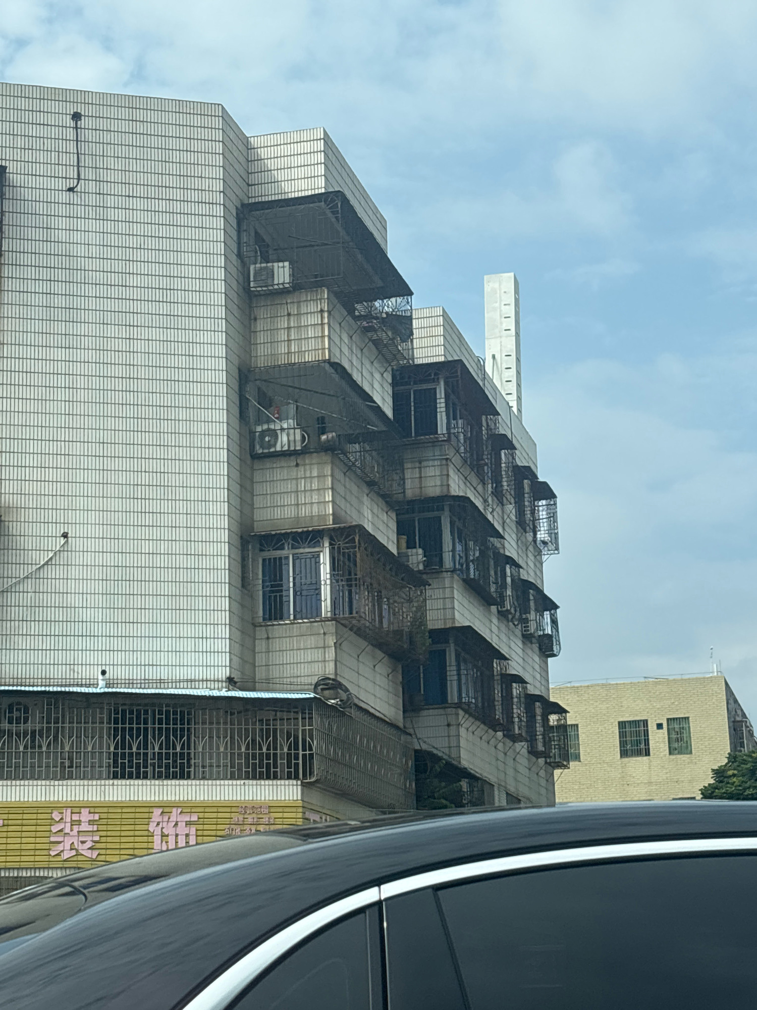 The image shows a tall, multi-story building with a light-colored, tiled facade in Sanshui District, China. The building has numerous balconies with metal railings and awnings, and several windows are visible, some with blue shutters. Air conditioning units are mounted on the exterior walls. To the right of the main building, a shorter, cream-colored building with multiple windows is visible. A black car with a silver trim is in the foreground, partially obscuring the lower part of the buildings and a yellow sign with pink Chinese characters. The sky is cloudy.